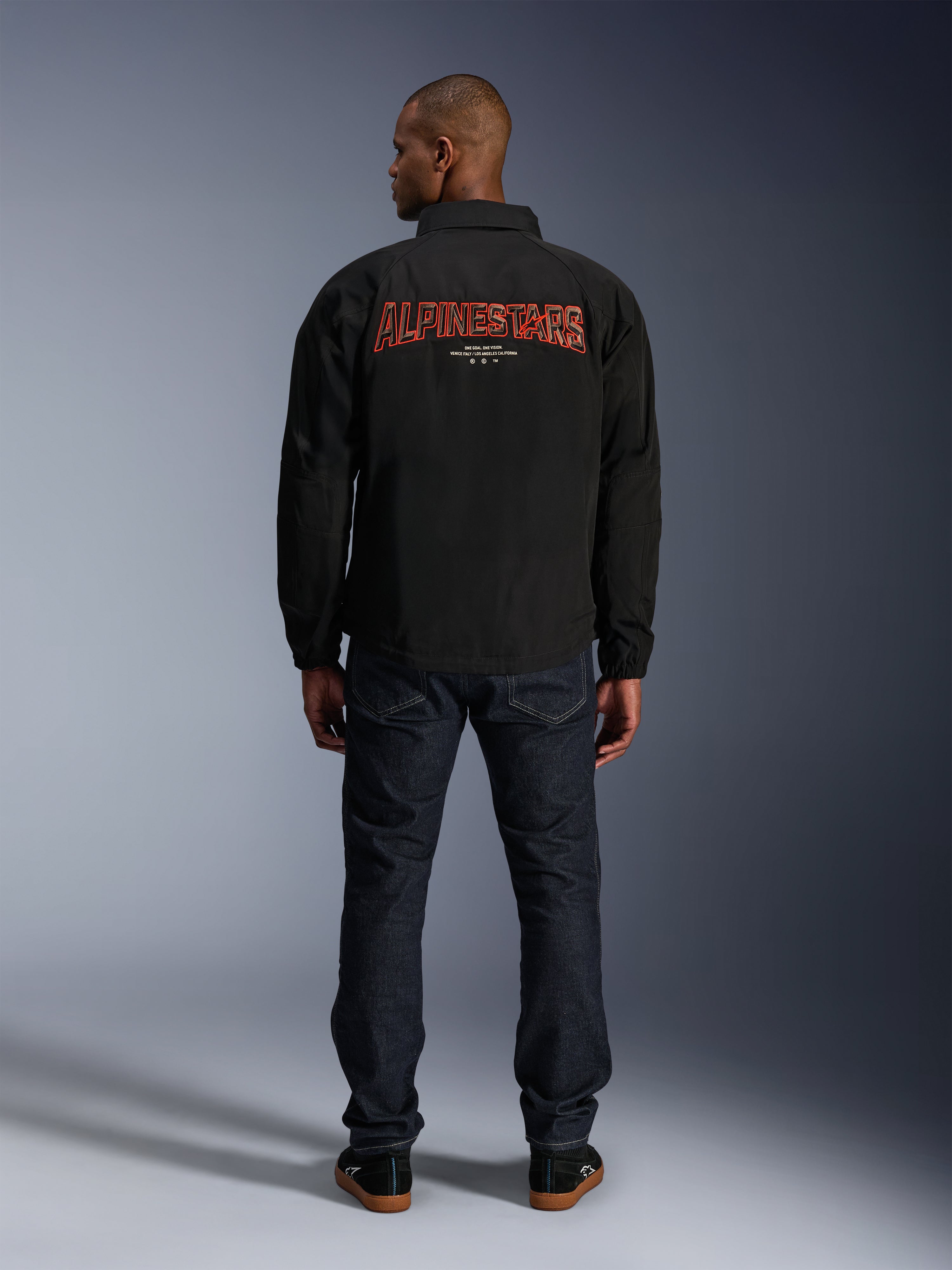 Alpinestars Revved WR Jacket, Black, back view featuring large red 3D-effect 'ALPINESTARS' embroidery, urban coaches-style jacket, worn with dark denim jeans and black sneakers, minimalist motorcycle apparel
