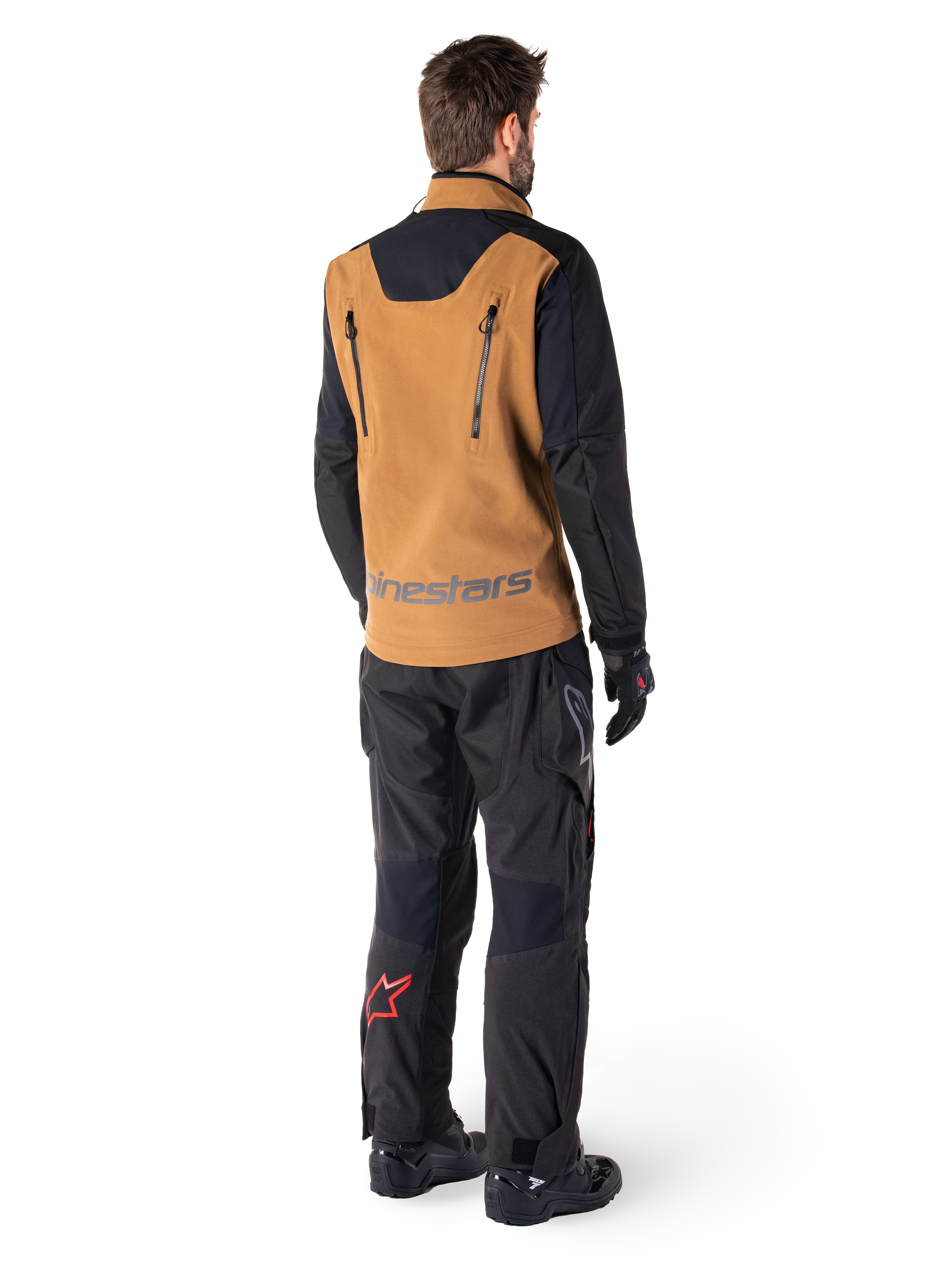 Casaco Alpinestars Hyde XT Stretch Drystar®XF e Calças Hyde XT Drystar®, vista traseira de um piloto com equipamento de adventure touring bege e preto, casaco com fechos verticais nas costas e marca Alpinestars, calças pretas com logótipo de estrela vermelha na barriga da perna, combinado com luvas e botas pretas.