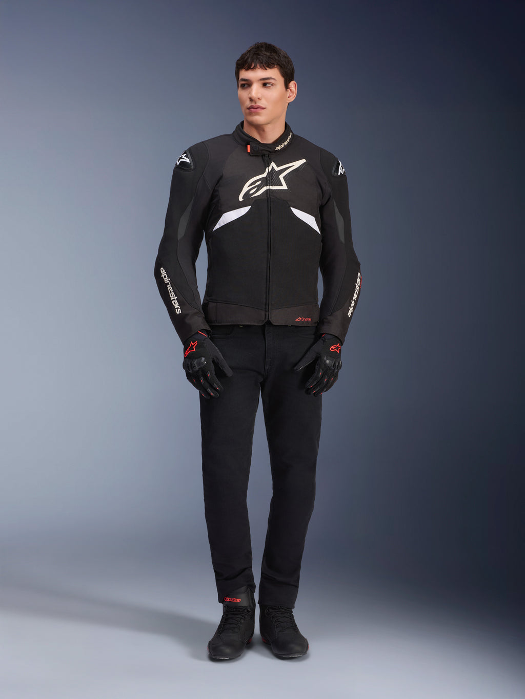 Modelo masculino a usar o Casaco Alpinestars T-GP R V3 Drystar® em preto e branco, casaco desportivo têxtil impermeável com um grande logótipo Alpinestars branco no peito, usado com calças e luvas de condução pretas, retrato de estúdio profissional realçando a silhueta desportiva elegante.
