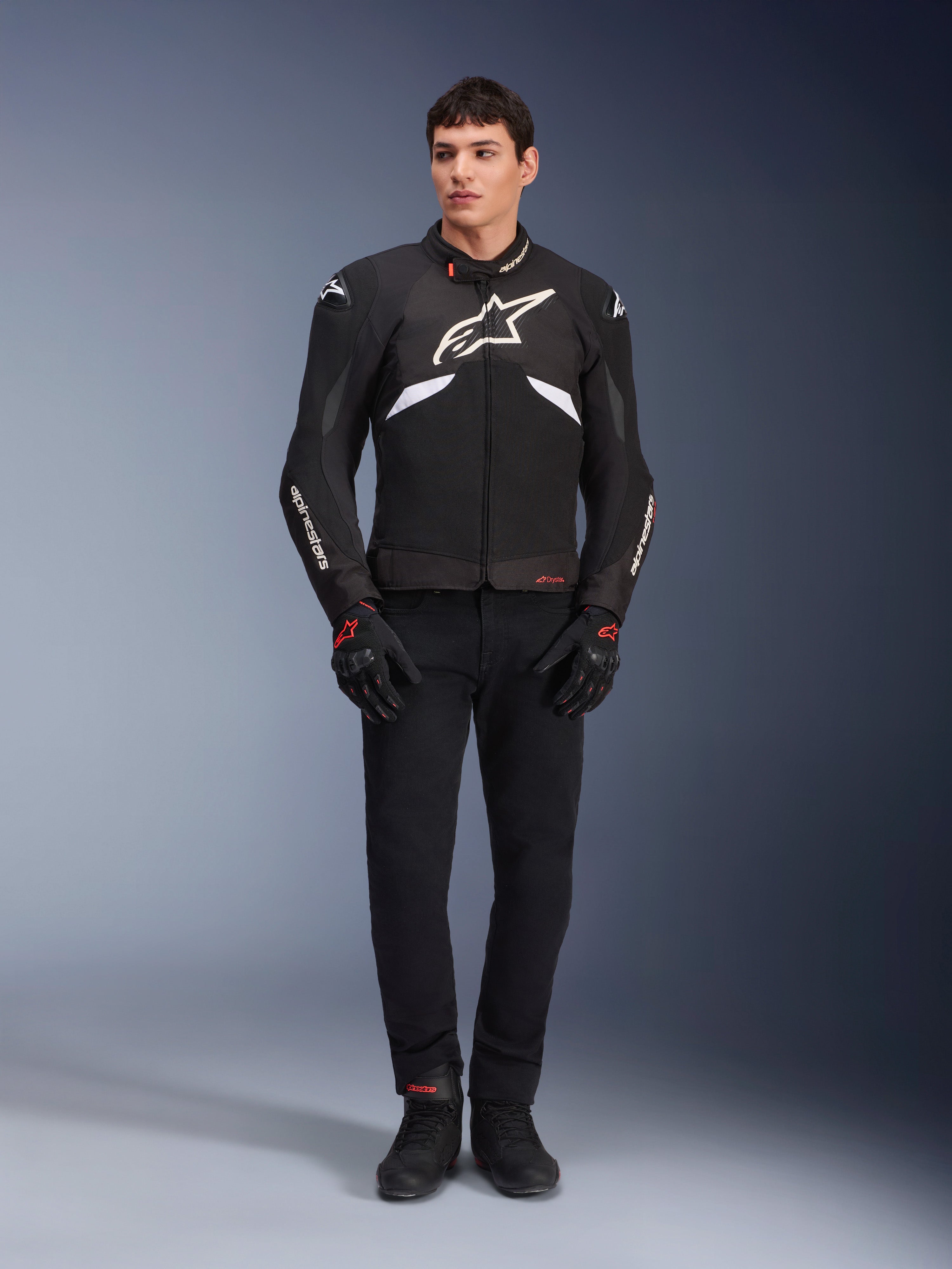 Modelo masculino a usar o Casaco Alpinestars T-GP R V3 Drystar® em preto e branco, casaco desportivo têxtil impermeável com um grande logótipo Alpinestars branco no peito, usado com calças e luvas de condução pretas, retrato de estúdio profissional realçando a silhueta desportiva elegante.