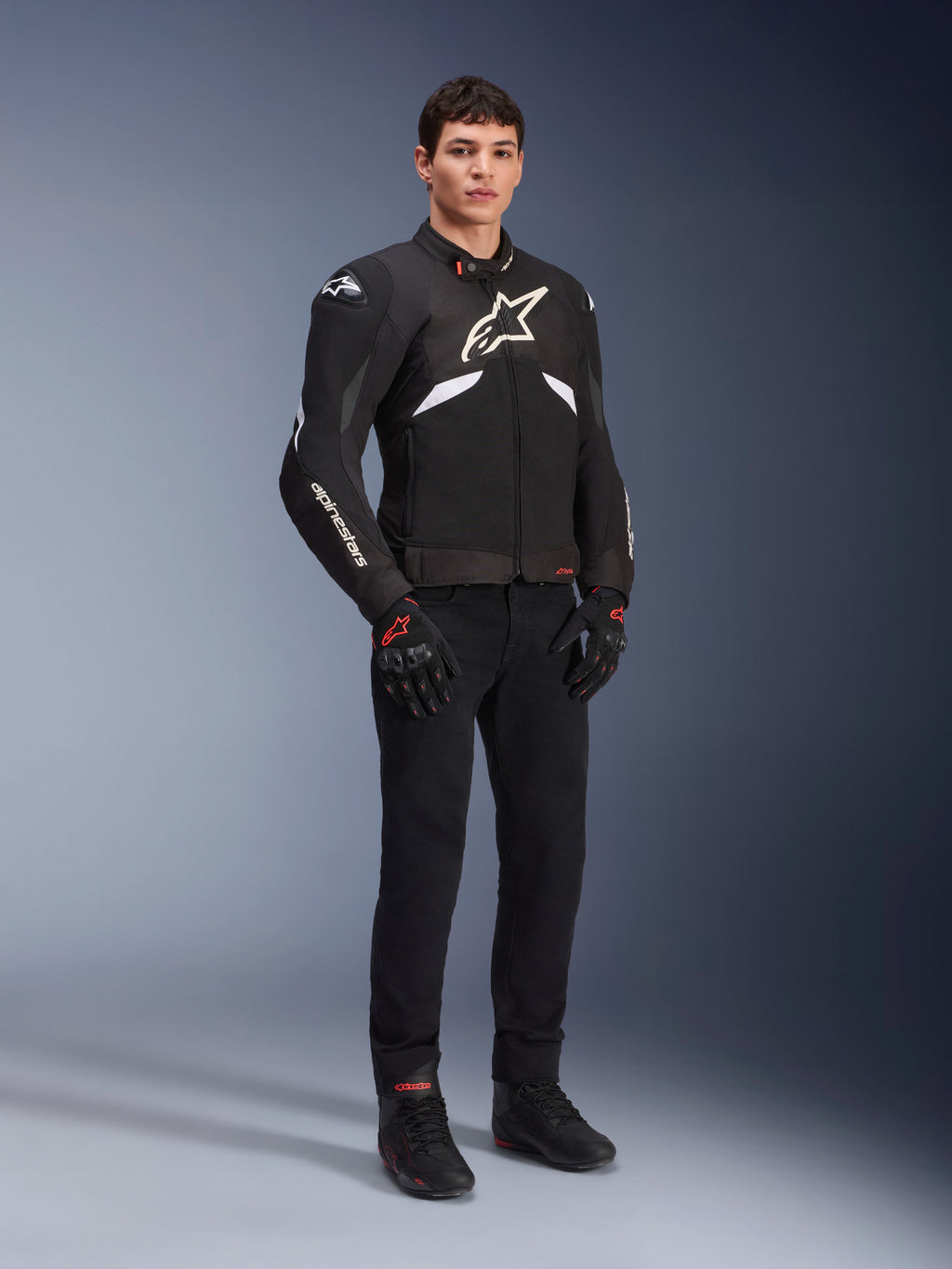 Casaco Alpinestars T-GP R V3 Drystar® em preto e branco, vestido por um modelo com luvas e calças de motociclismo pretas, com logótipo branco no peito, detalhes brancos nas mangas e deslizadores de ombro protetores, casaco desportivo têxtil impermeável para condução em estrada