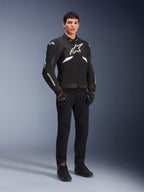 Casaco Alpinestars T-GP R V3 Drystar® em preto e branco, vestido por um modelo com luvas e calças de motociclismo pretas, com logótipo branco no peito, detalhes brancos nas mangas e deslizadores de ombro protetores, casaco desportivo têxtil impermeável para condução em estrada