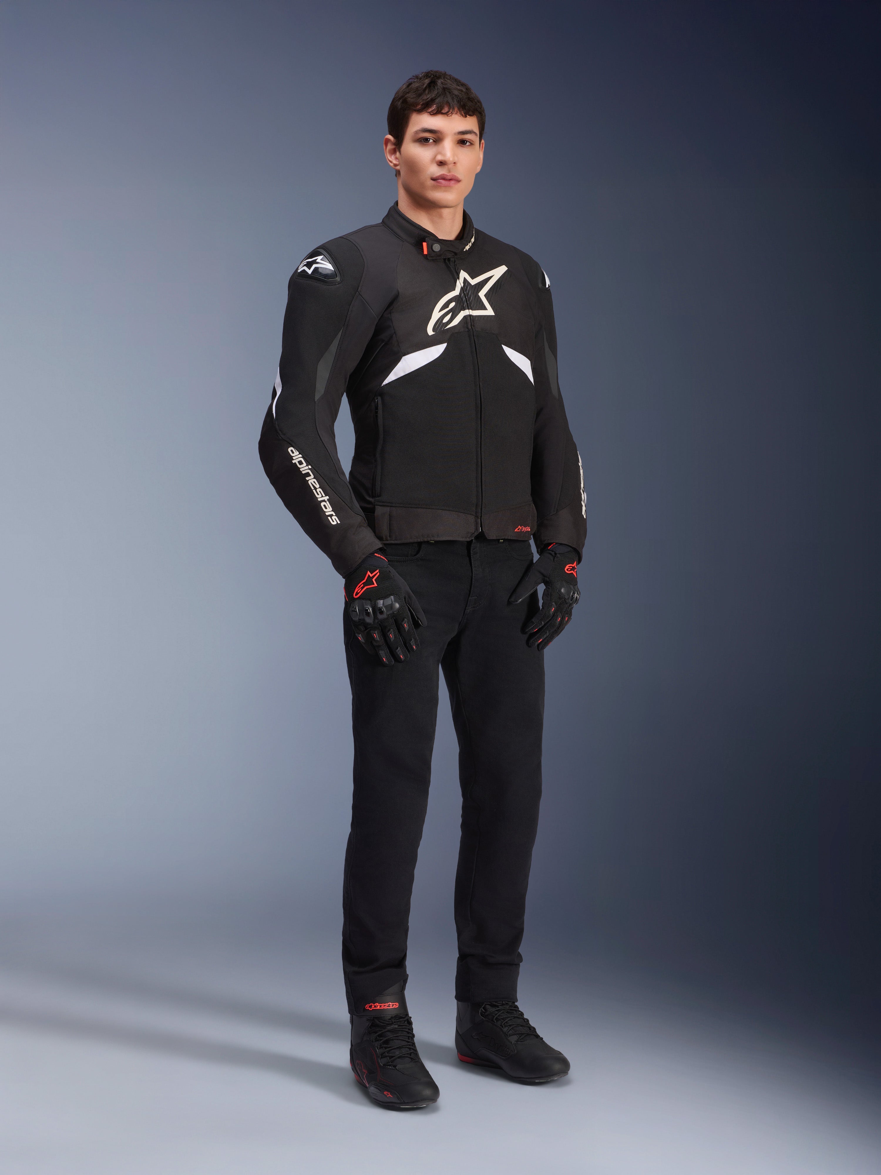 Casaco Alpinestars T-GP R V3 Drystar® em preto e branco, vestido por um modelo com luvas e calças de motociclismo pretas, com logótipo branco no peito, detalhes brancos nas mangas e deslizadores de ombro protetores, casaco desportivo têxtil impermeável para condução em estrada