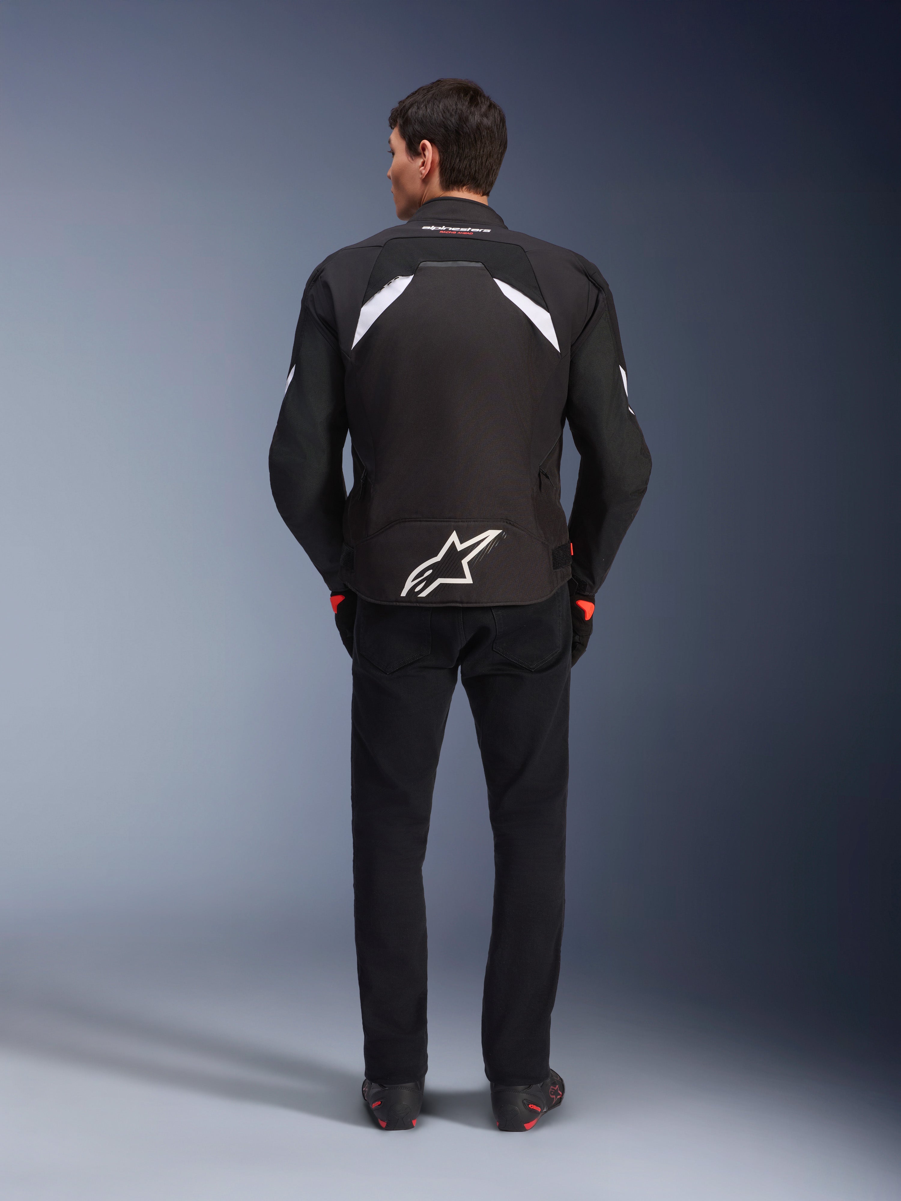 Casaco Alpinestars T-GP R V3 Drystar®, preto e branco, vista traseira, casaco têxtil de motociclismo impermeável com um logótipo de estrela branca grande na parte inferior das costas e detalhes brancos nos ombros, combinado com calças e botas pretas, design desportivo derivado da competição