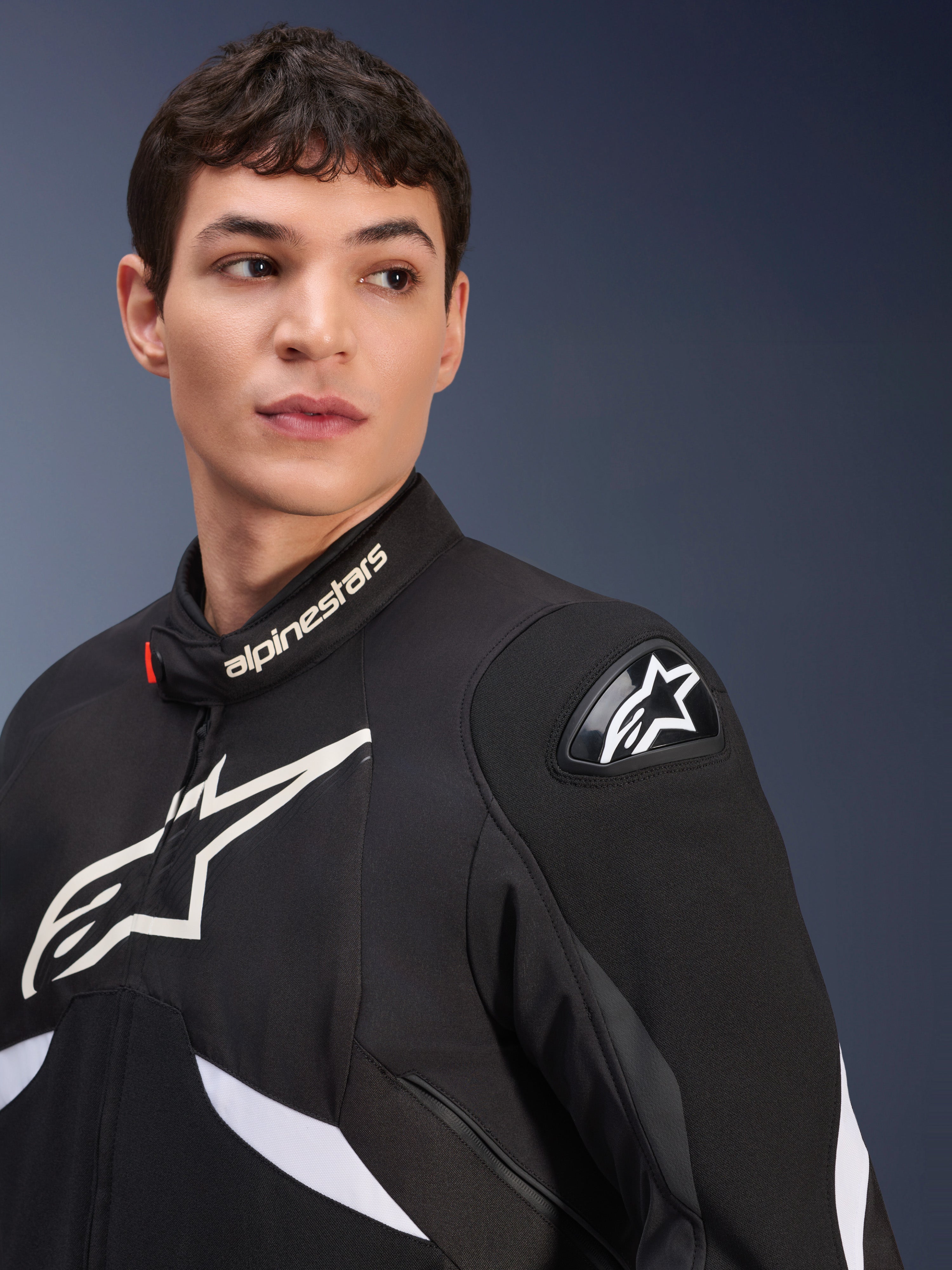 Casaco Alpinestars T-GP R V3 Drystar®, preto e branco, vestido por um modelo masculino, com logótipos Alpinestars brancos na gola e no peito, deslizador de ombro DFS preto com uma estrela branca, imagem detalhada de estilo de vida de um casaco desportivo têxtil impermeável