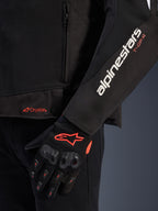 Casaco Alpinestars T-GP R V3 Drystar®, casaco de motociclismo têxtil preto, vista aproximada mostrando a marca Alpinestars branca e T-GP R laranja na manga, logótipo Drystar laranja na bainha, acompanhado por luvas de malha protetoras pretas com detalhes vermelhos.