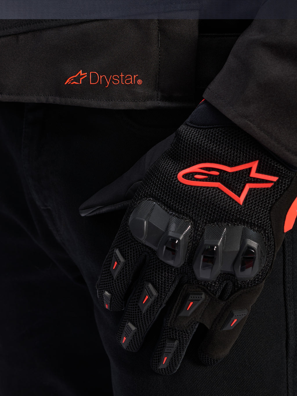 Pormenor da cintura do casaco Alpinestars T-GP R V3 Drystar® com logótipo Drystar vermelho, mostrado com uma luva de motociclismo preta com logótipo de estrela Alpinestars vermelho, tecido em malha e proteção reforçada nos nós dos dedos.