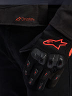 Pormenor da cintura do casaco Alpinestars T-GP R V3 Drystar® com logótipo Drystar vermelho, mostrado com uma luva de motociclismo preta com logótipo de estrela Alpinestars vermelho, tecido em malha e proteção reforçada nos nós dos dedos.