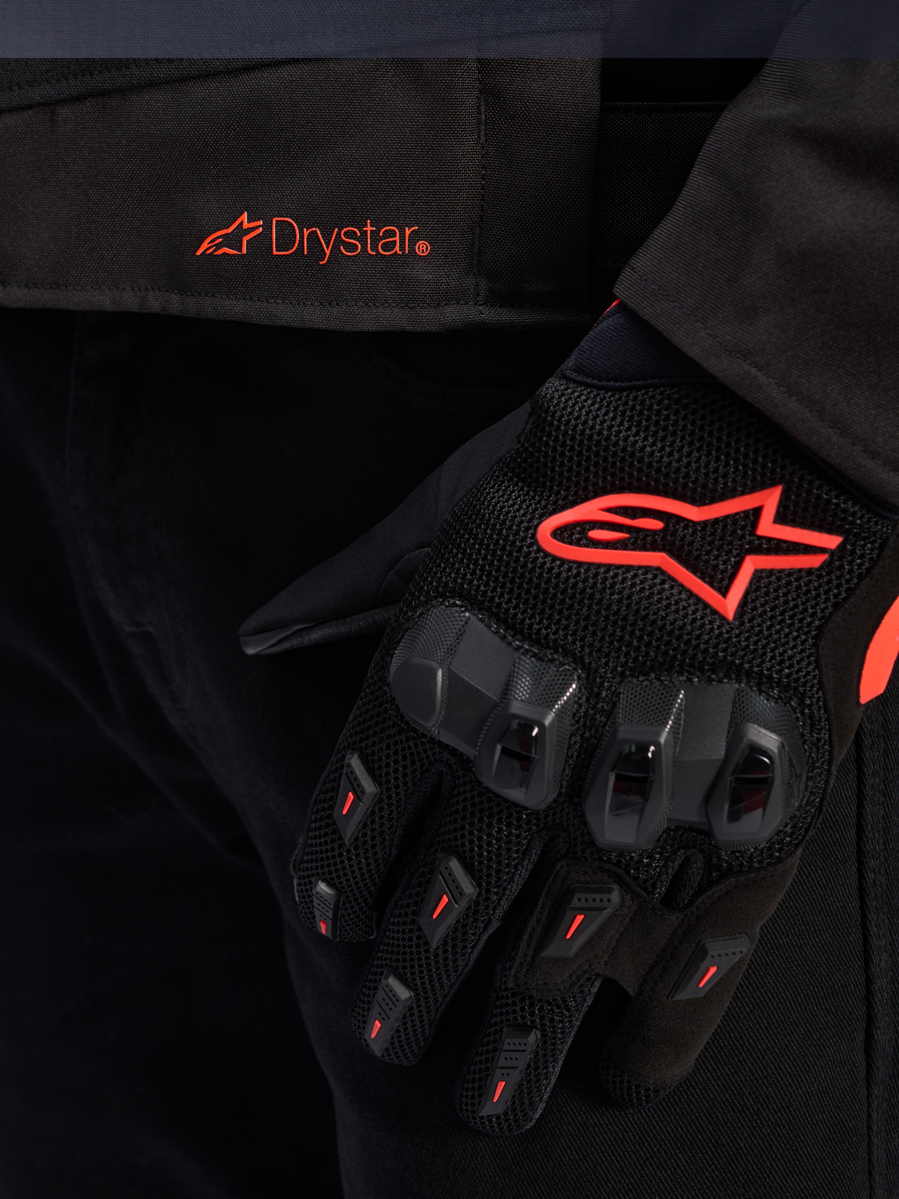 Pormenor da cintura do casaco Alpinestars T-GP R V3 Drystar® com logótipo Drystar vermelho, mostrado com uma luva de motociclismo preta com logótipo de estrela Alpinestars vermelho, tecido em malha e proteção reforçada nos nós dos dedos.
