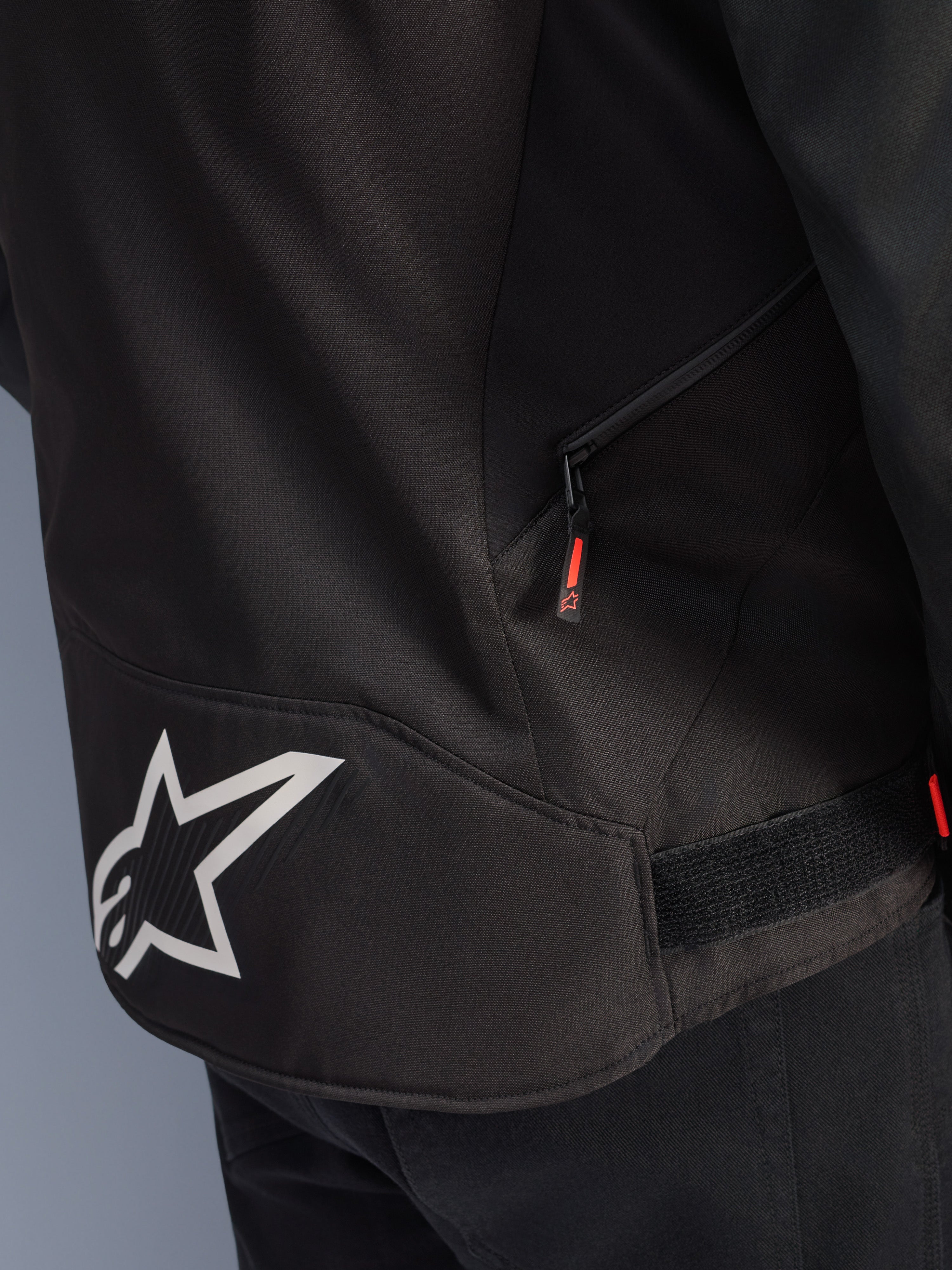 Casaco Alpinestars T-GP R V3 Drystar®, preto e branco, vista de pormenor da zona lombar com logótipo de estrela Alpinestars grande em branco, bolso lateral com fecho e puxador vermelho, tira de ajuste de cintura em velcro, material têxtil resistente