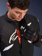Casaco Alpinestars T-GP R V3 Drystar®, casaco têxtil desportivo de motociclismo em preto e branco, parcialmente aberto revelando fita interior cor de laranja 'Alpinestars Protects', usado com luvas de malha pretas e vermelhas, vestuário de proteção derivado da competição.