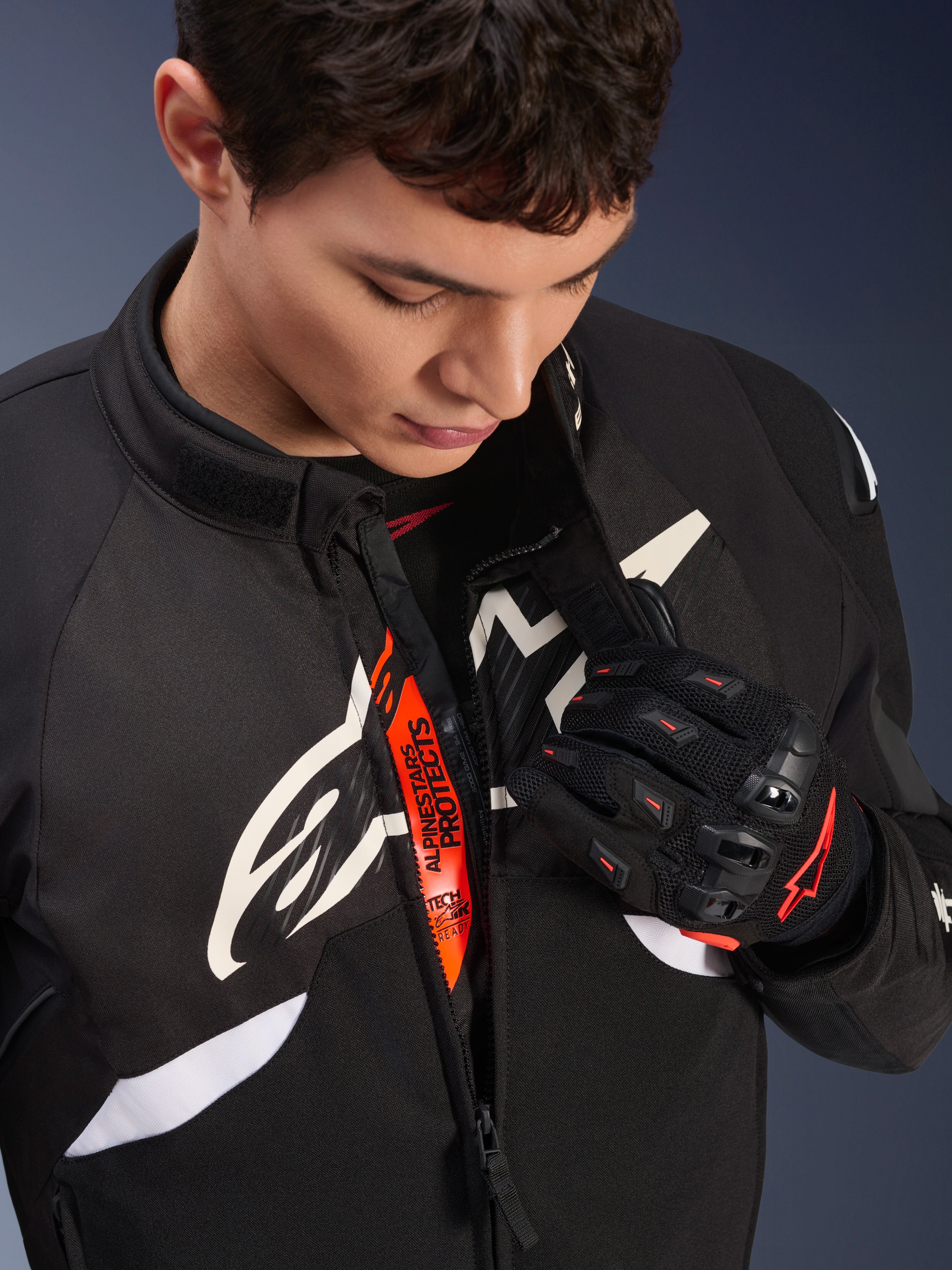 Casaco Alpinestars T-GP R V3 Drystar®, casaco têxtil desportivo de motociclismo em preto e branco, parcialmente aberto revelando fita interior cor de laranja 'Alpinestars Protects', usado com luvas de malha pretas e vermelhas, vestuário de proteção derivado da competição.
