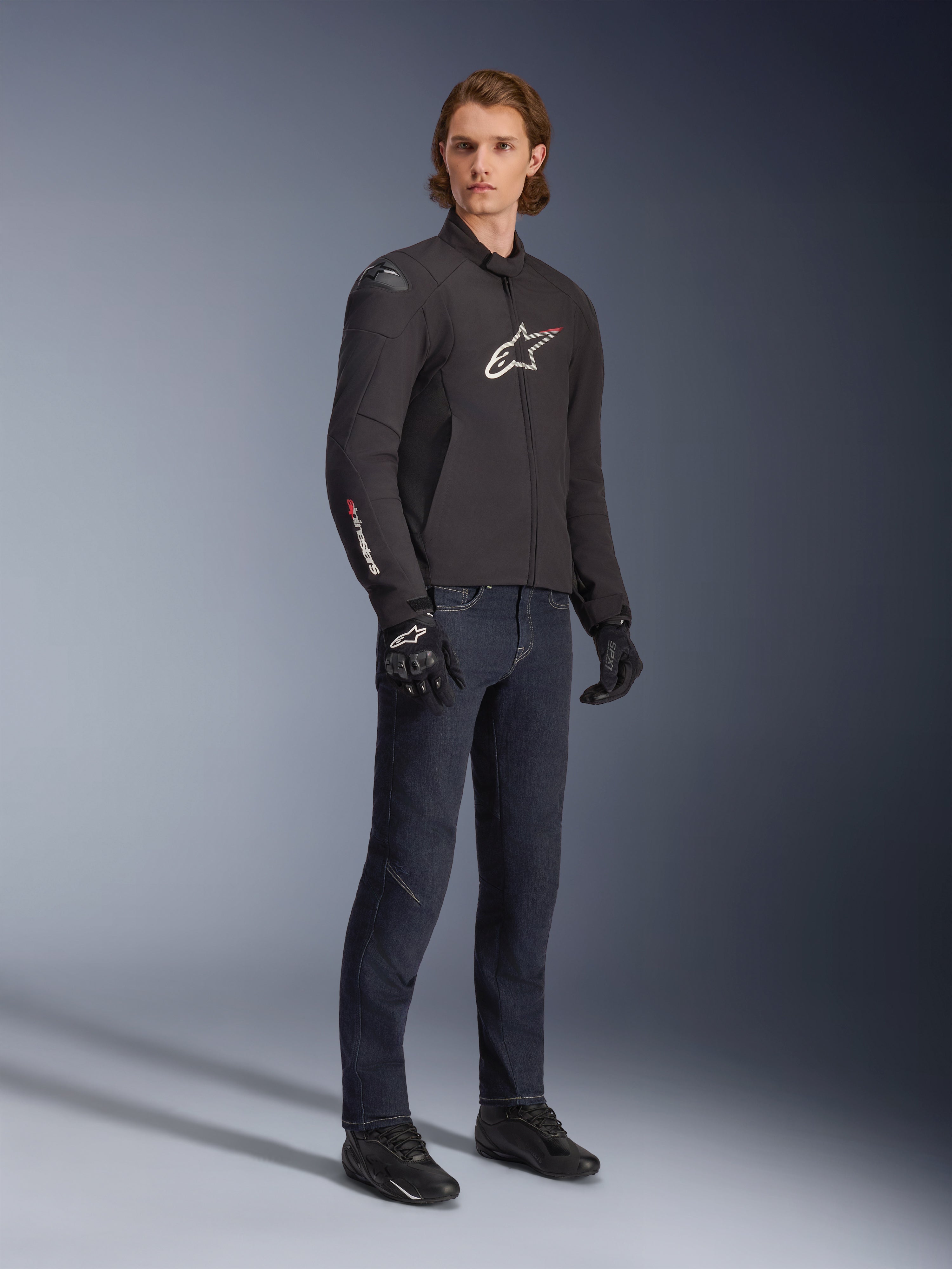 Male model wearing Alpinestars SMX Waterproof Jacket, Black with white and red logo accents, styled with dark blue denim pants and black riding gloves, sleek sport-fit textile motorcycle jacket.