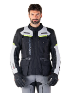 Homem a usar o casaco Alpinestars Bogotá Pro Drystar®, preto, cinzento e amarelo néon, casaco de motociclismo adventure touring para todas as estações, design impermeável com múltiplos bolsos e marca vertical, combinado com luvas pretas e calças de condução cinzentas
