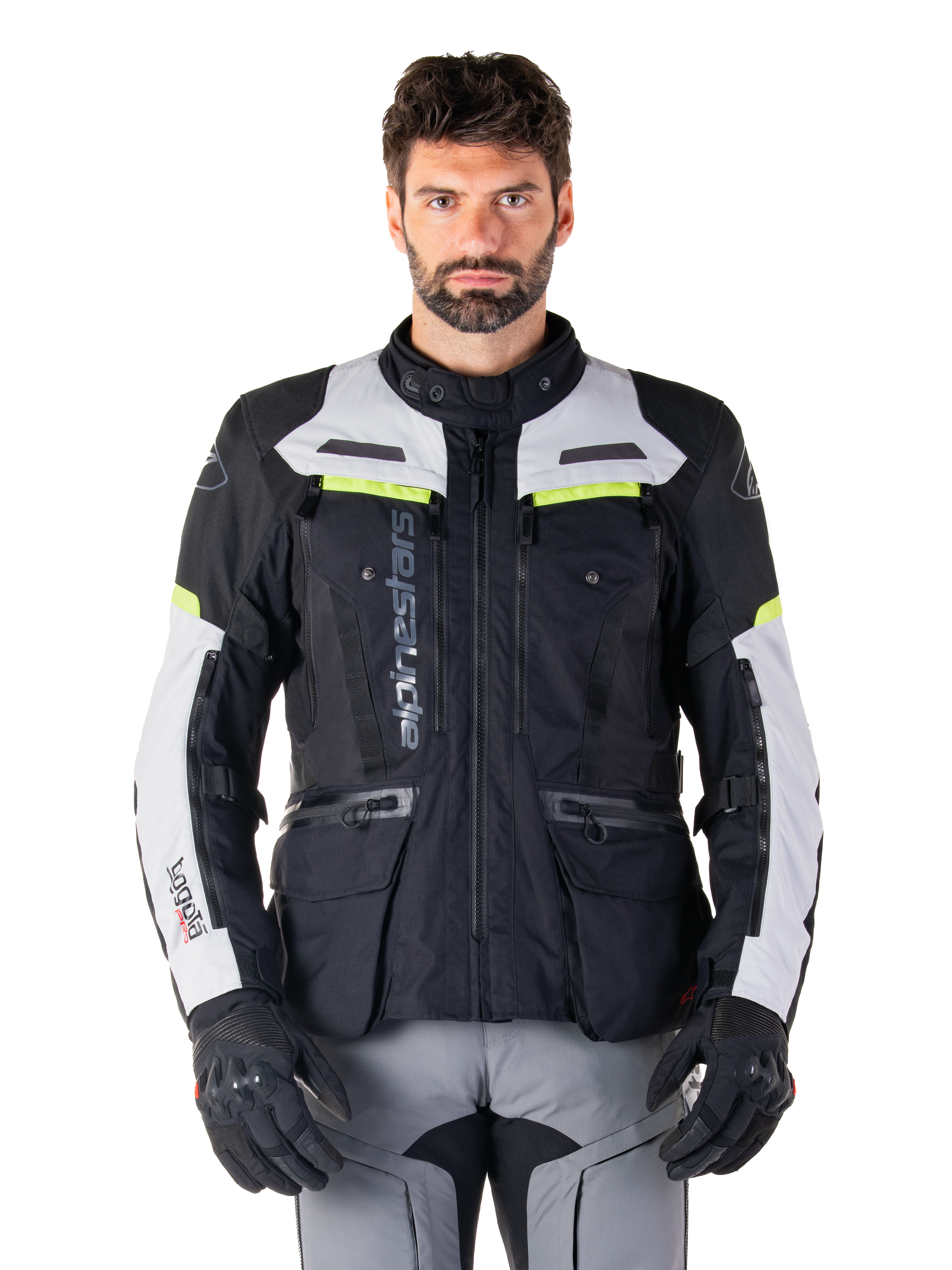 Homem a usar o casaco Alpinestars Bogotá Pro Drystar®, preto, cinzento e amarelo néon, casaco de motociclismo adventure touring para todas as estações, design impermeável com múltiplos bolsos e marca vertical, combinado com luvas pretas e calças de condução cinzentas