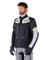 Casaco Alpinestars Bogotá Pro Drystar®, casaco de motociclismo adventure touring, preto, cinzento gelo e amarelo fluo, usado por um homem com calças cinzentas a condizer e luvas pretas, com painéis de ventilação técnicos e design com vários bolsos.