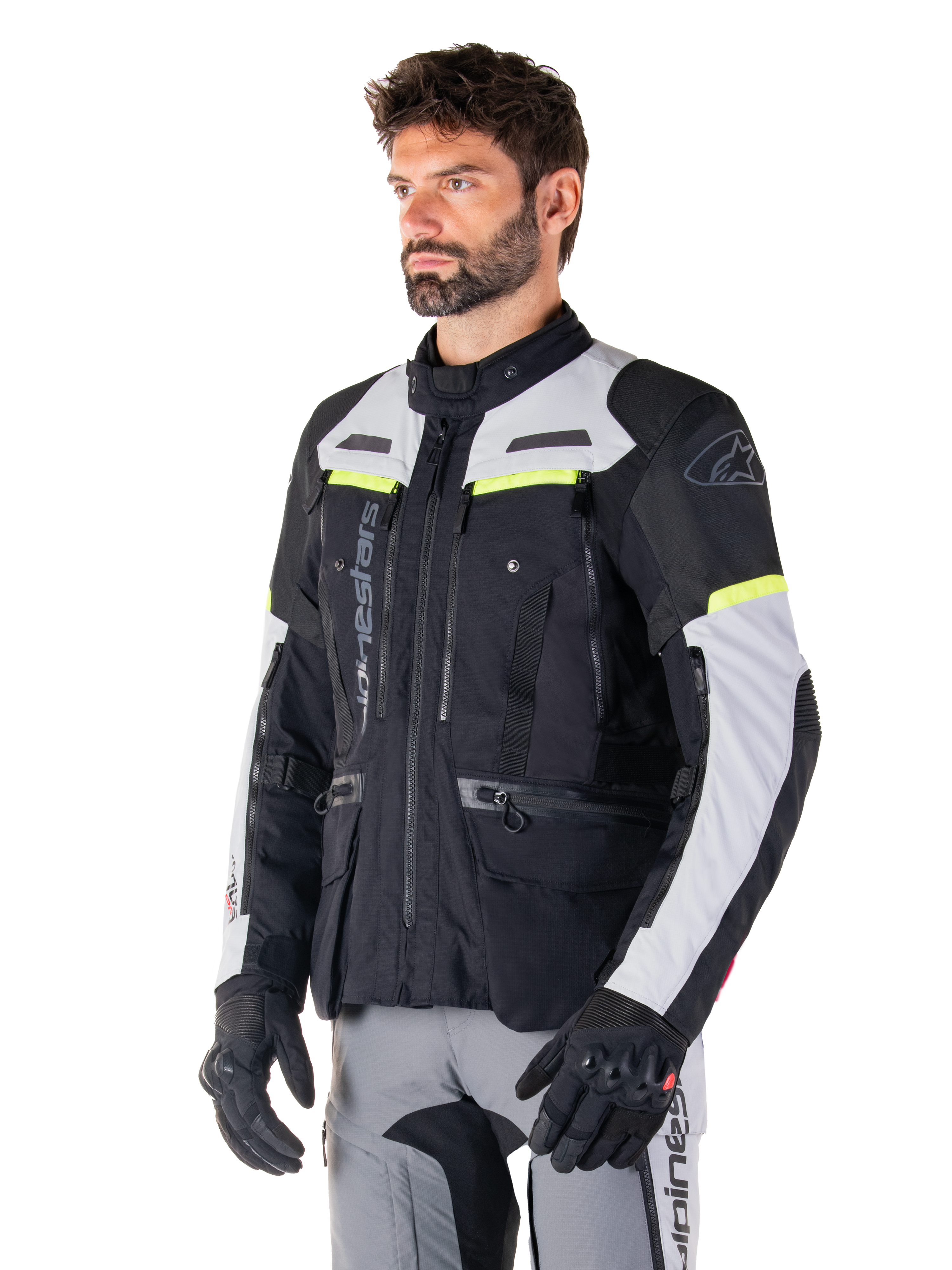 Casaco Alpinestars Bogotá Pro Drystar®, casaco de motociclismo adventure touring, preto, cinzento gelo e amarelo fluo, usado por um homem com calças cinzentas a condizer e luvas pretas, com painéis de ventilação técnicos e design com vários bolsos.