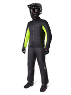 Motociclista a usar o forro do casaco Alpinestars Bogotá Pro Drystar®, preto com painéis em amarelo néon brilhante nas mangas, usado com calças pretas a condizer, capacete de motocross preto e luvas, equipamento de aventura impermeável para todas as estações