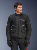 Casaco Alpinestars Andes Air Drystar®, casaco de motociclismo touring preto, logótipo de estrela vermelha no peito, com painéis de ventilação em malha e detalhes refletores, usado por modelo masculino com luvas de condução pretas, equipamento de aventura para duas estações