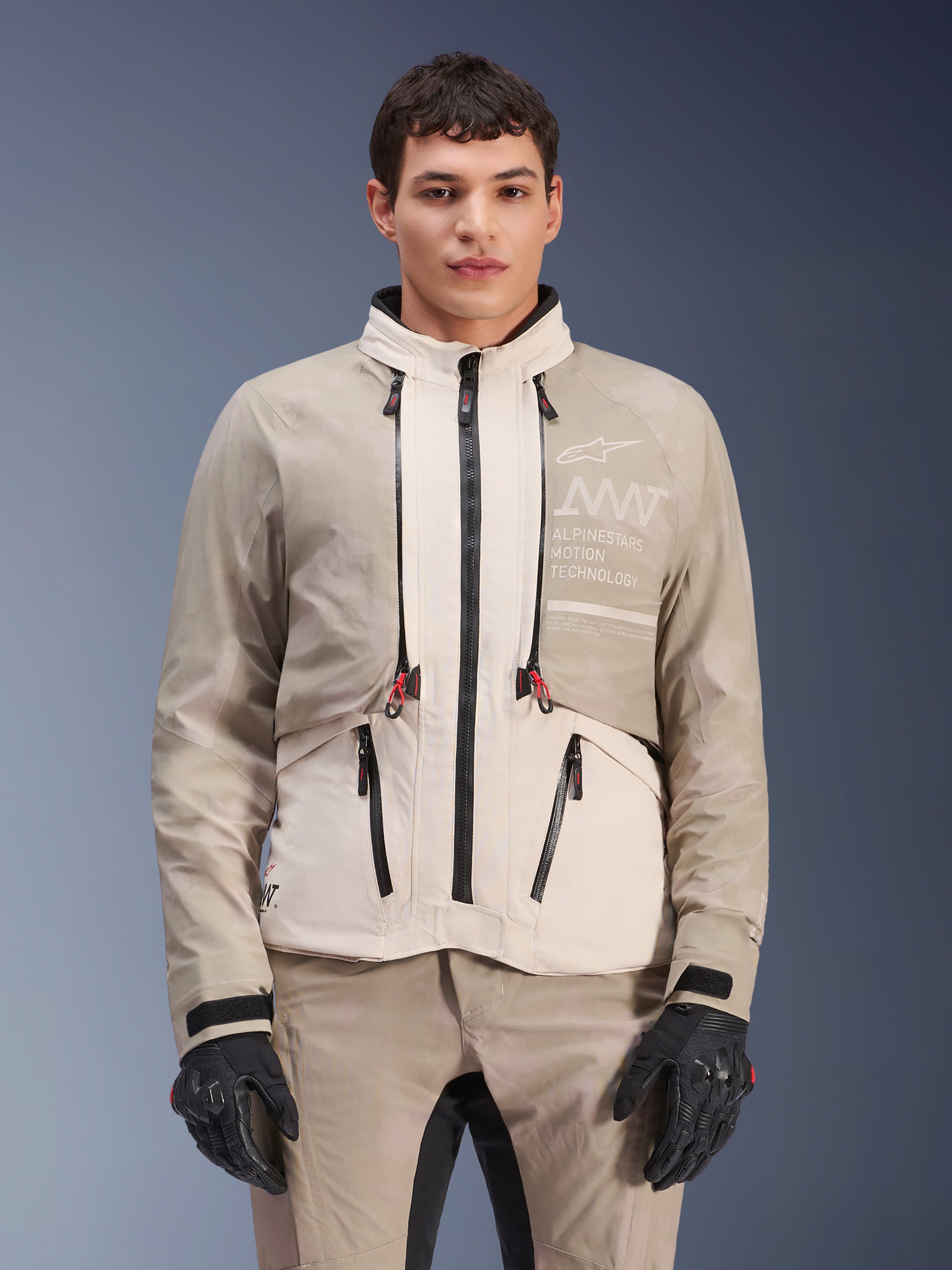 Casaco Alpinestars AMT-10LAB Drystar® XF, casaco de motociclismo de aventura, White Sand Aluminum Camo, design modular com fechos pretos e pormenores vermelhos, usado com calças coordenadas e luvas pretas, com o logótipo Alpinestars Motion Technology no peito, fato de condução profissional para todas as condições meteorológicas