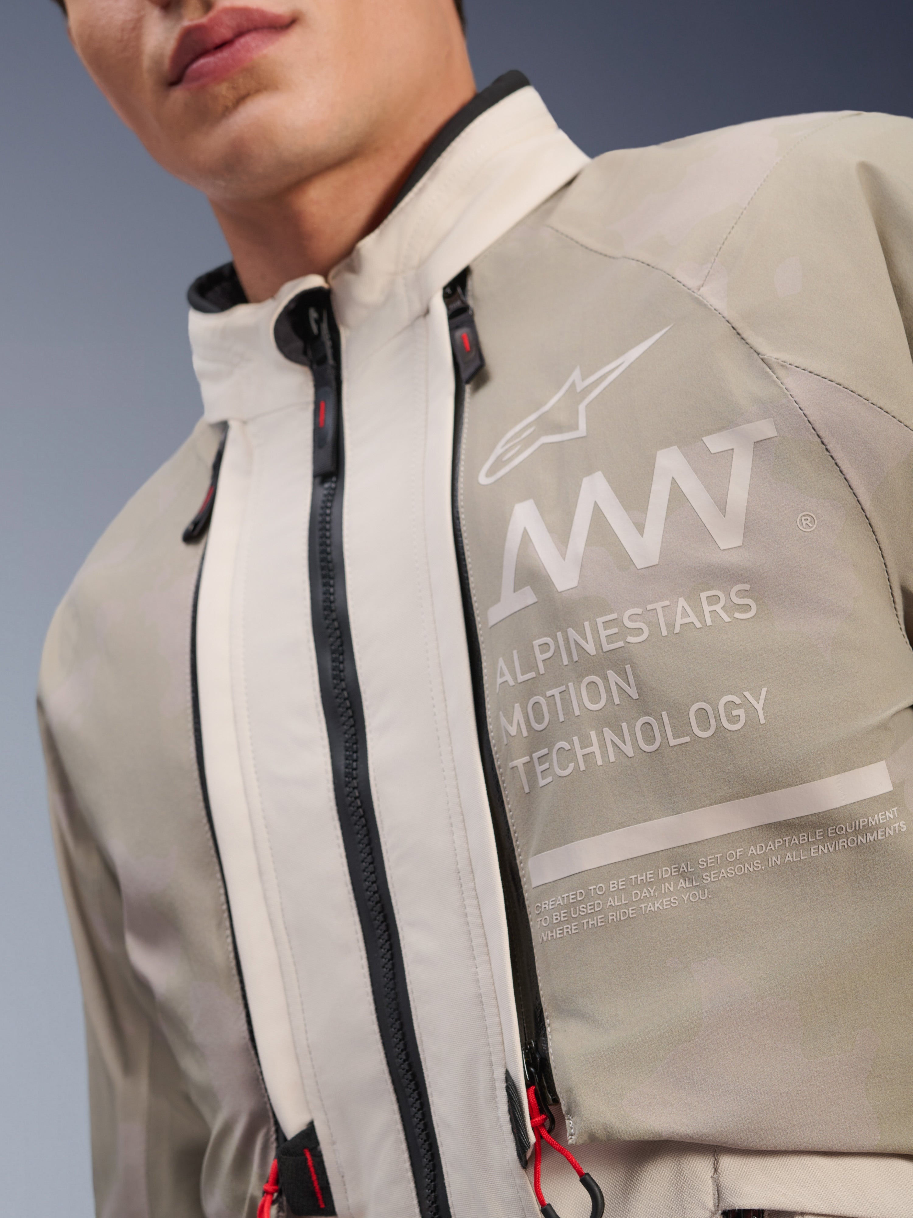 Casaco Alpinestars AMT-10LAB Drystar® XF, White Sand Aluminum Camo, detalhe do peito com a marca AMT Alpinestars Motion Technology, fechos de correr impermeáveis com puxadores vermelhos e tecido têxtil texturizado preparado para aventura.