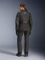 Condutora vista de trás a usar o Casaco Alpinestars Stella Andes V4 Drystar®, preto, combinado com calças de motociclismo a condizer, logótipo Astar branco nas costas, bainha traseira alongada, equipamento completo de condução adventure-touring, incluindo luvas e botas pretas