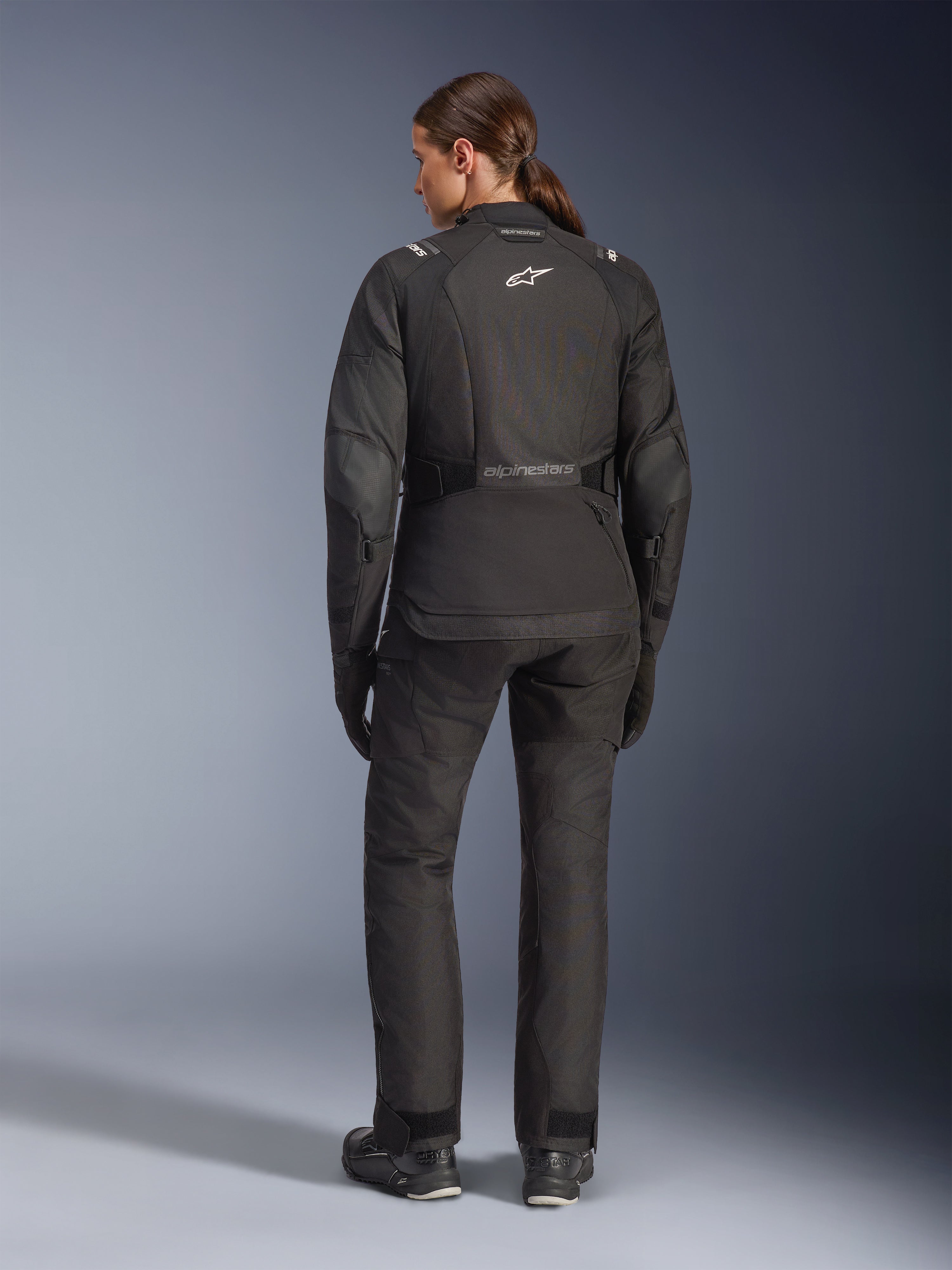 Condutora vista de trás a usar o Casaco Alpinestars Stella Andes V4 Drystar®, preto, combinado com calças de motociclismo a condizer, logótipo Astar branco nas costas, bainha traseira alongada, equipamento completo de condução adventure-touring, incluindo luvas e botas pretas