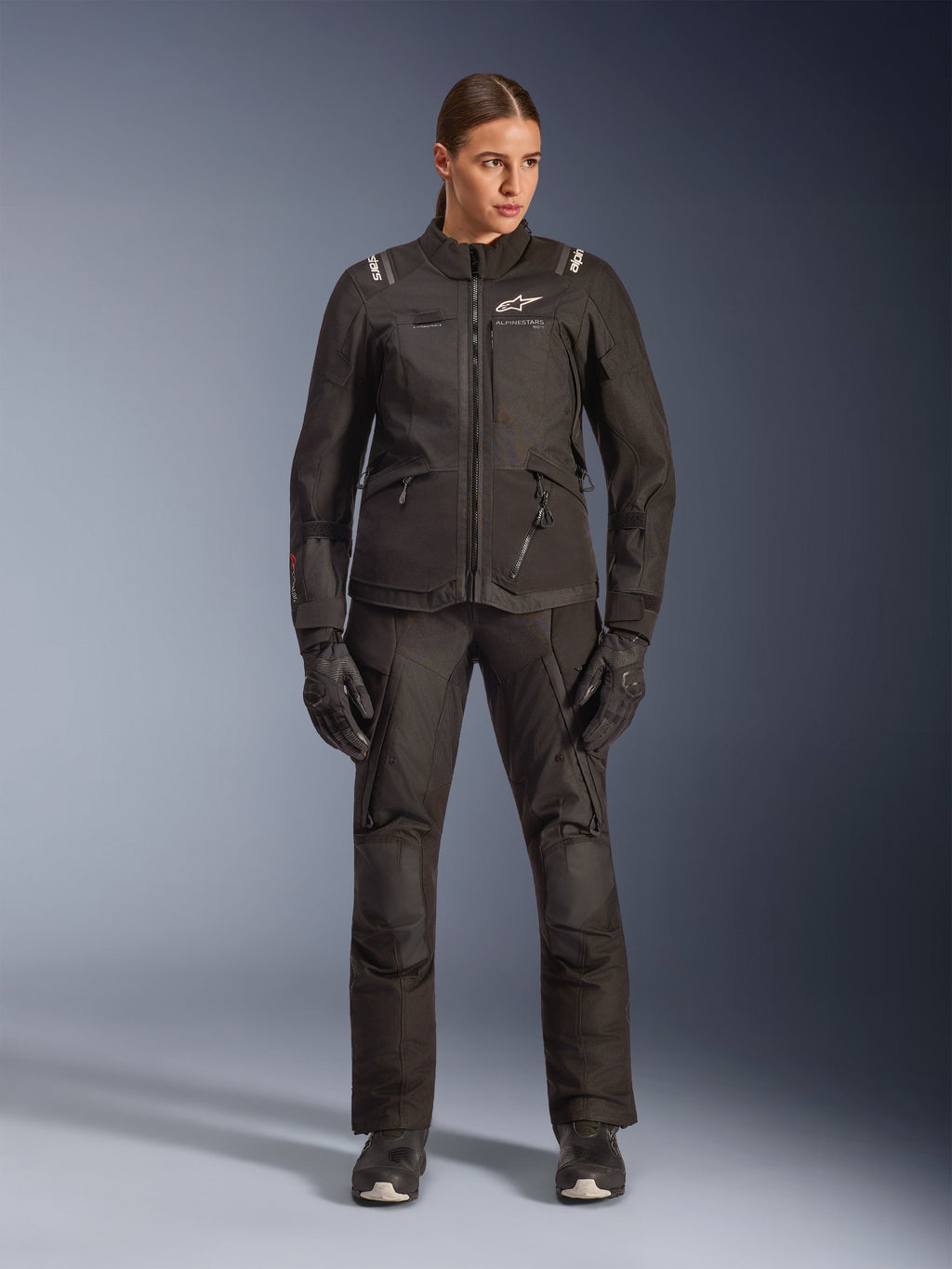 Motociclista feminina com o casaco Alpinestars Stella Andes V4 Drystar® em preto, combinado com calças e luvas de adventure-touring pretas a condizer, construção em têxtil técnico com múltiplos bolsos utilitários, logótipo Alpinestars em branco, equipamento de motociclismo completo para todas as condições climatéricas