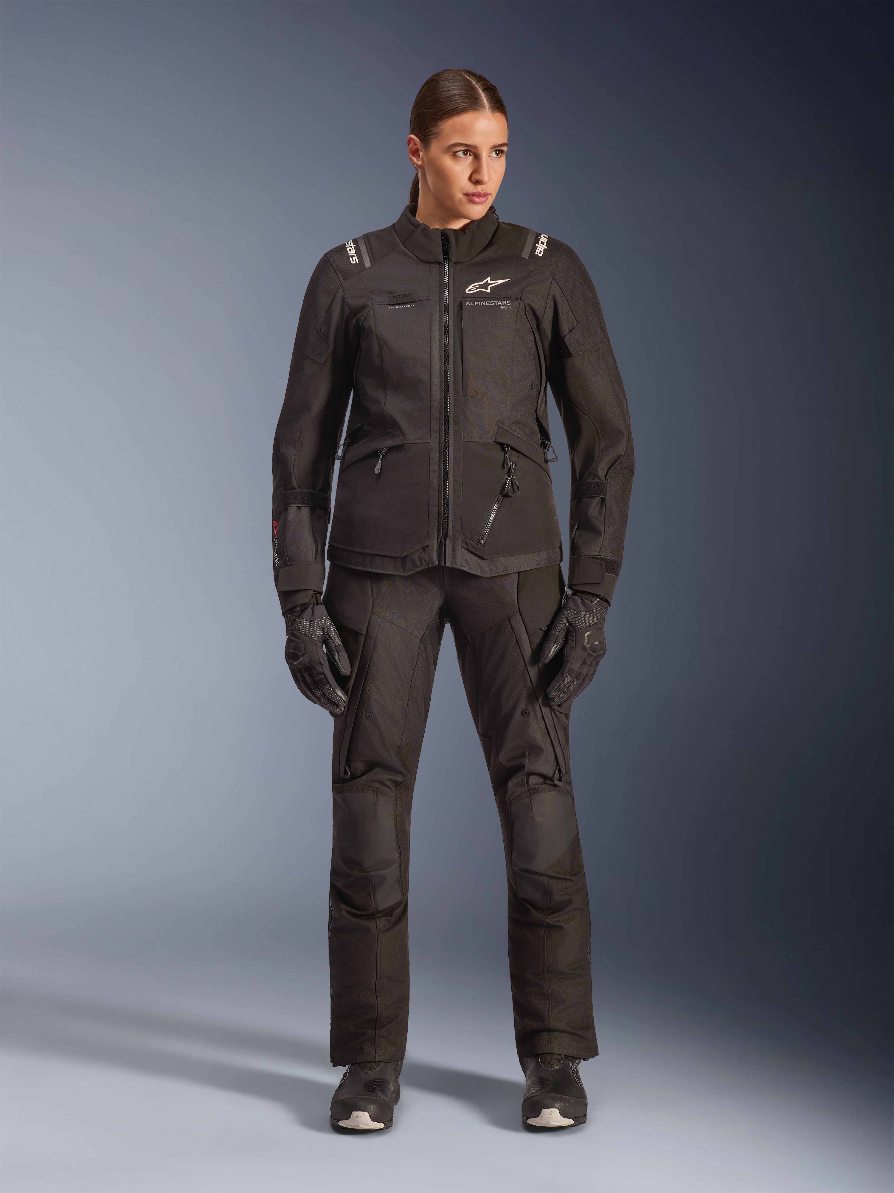 Motociclista feminina com o casaco Alpinestars Stella Andes V4 Drystar® em preto, combinado com calças e luvas de adventure-touring pretas a condizer, construção em têxtil técnico com múltiplos bolsos utilitários, logótipo Alpinestars em branco, equipamento de motociclismo completo para todas as condições climatéricas