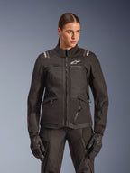 Casaco Alpinestars Stella Andes V4 Drystar, casaco de motociclismo de turismo para mulher, preto, usado por uma modelo com calças e luvas de motociclismo a condizer, com fecho de correr frontal, logótipo branco no peito e marca Alpinestars nos ombros, equipamento elegante para qualquer clima.