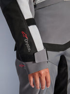 Alpinestars Stella Andes V4 Drystar®, casaco de aventura feminino cinzento, pormenor da manga com logótipo e fecho de velcro.