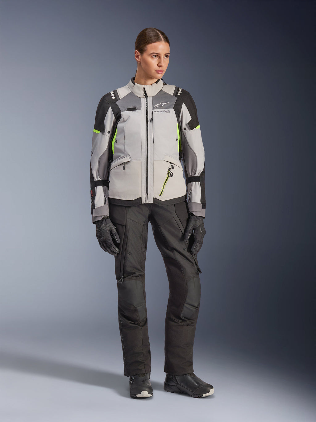 Modelo feminina a usar o Casaco Alpinestars Stella Andes V4 Drystar® em cinzento gelo, cinzento escuro e preto com pormenores em amarelo fluo, combinado com calças e luvas de motociclismo pretas, fotografia de estúdio de corpo inteiro de equipamento de aventura touring.