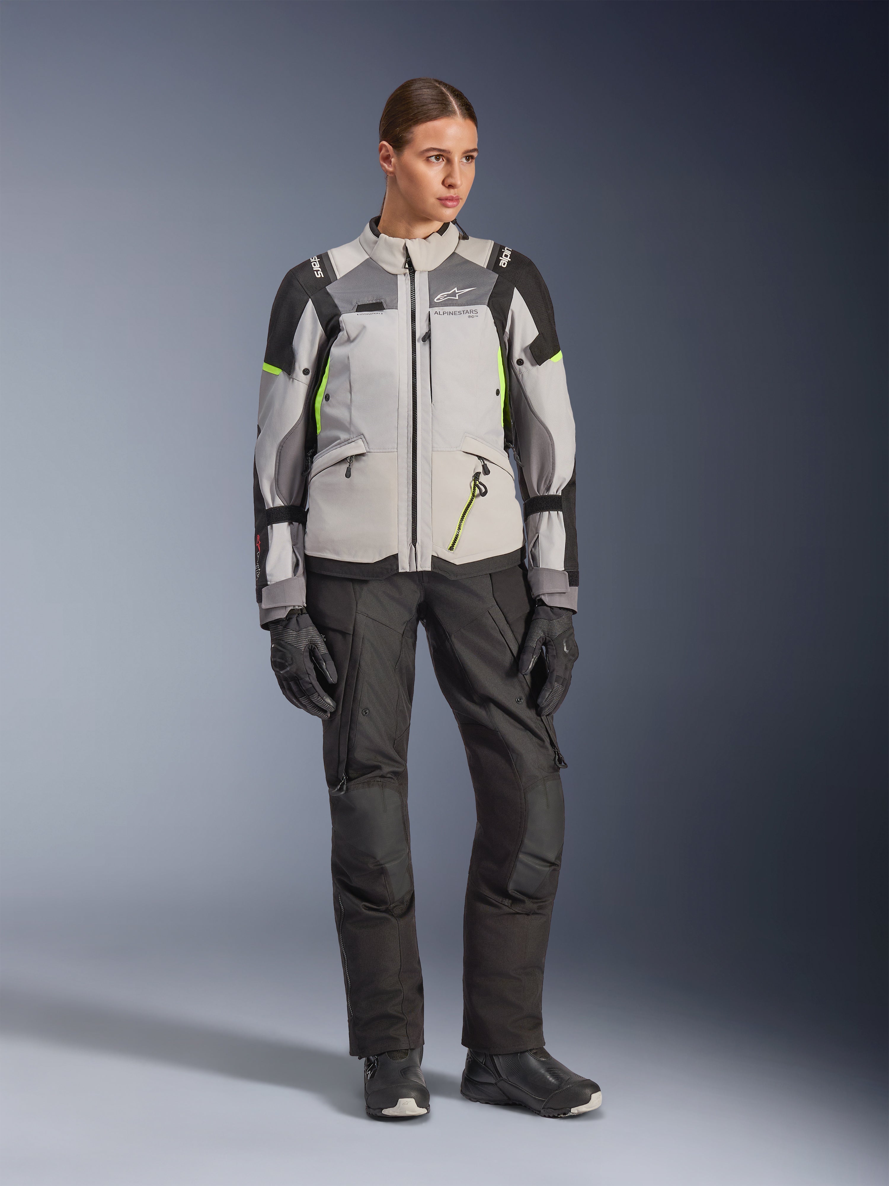 Modelo feminina a usar o Casaco Alpinestars Stella Andes V4 Drystar® em cinzento gelo, cinzento escuro e preto com pormenores em amarelo fluo, combinado com calças e luvas de motociclismo pretas, fotografia de estúdio de corpo inteiro de equipamento de aventura touring.