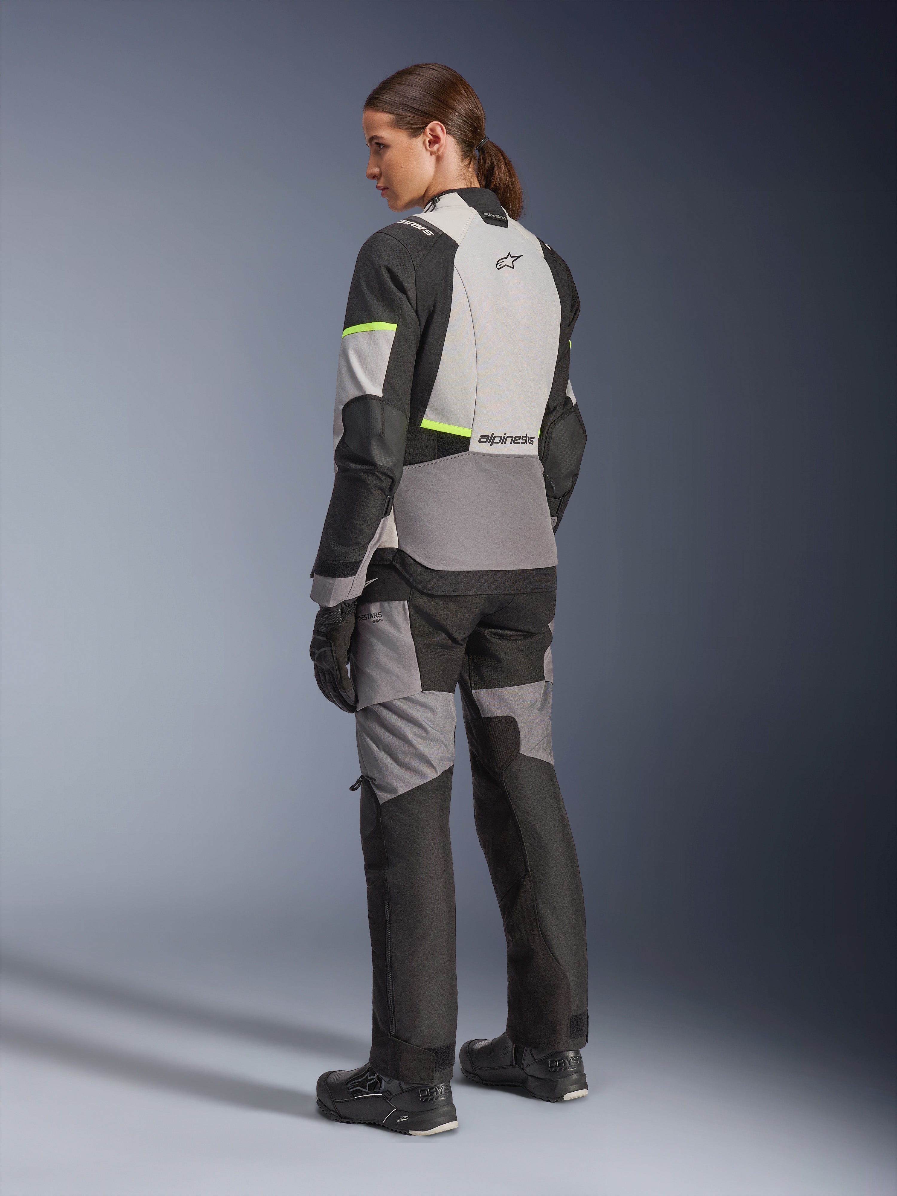 Casaco Alpinestars Stella Andes V4 Drystar® Feminino, casaco de motociclismo adventure-touring, cinzento gelo e cinzento escuro com detalhes em amarelo néon, vista traseira numa modelo a usar calças têxteis a condizer, com construção impermeável e painéis ripstop reforçados.