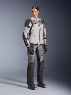 Mulher com casaco Alpinestars Stella Andes V4 Drystar® e calças a condizer, em Ice Gray e Dark Gray com detalhes em amarelo néon, conjunto completo de equipamento de aventura touring com luvas e botas pretas, fotografia de estúdio de corpo inteiro.