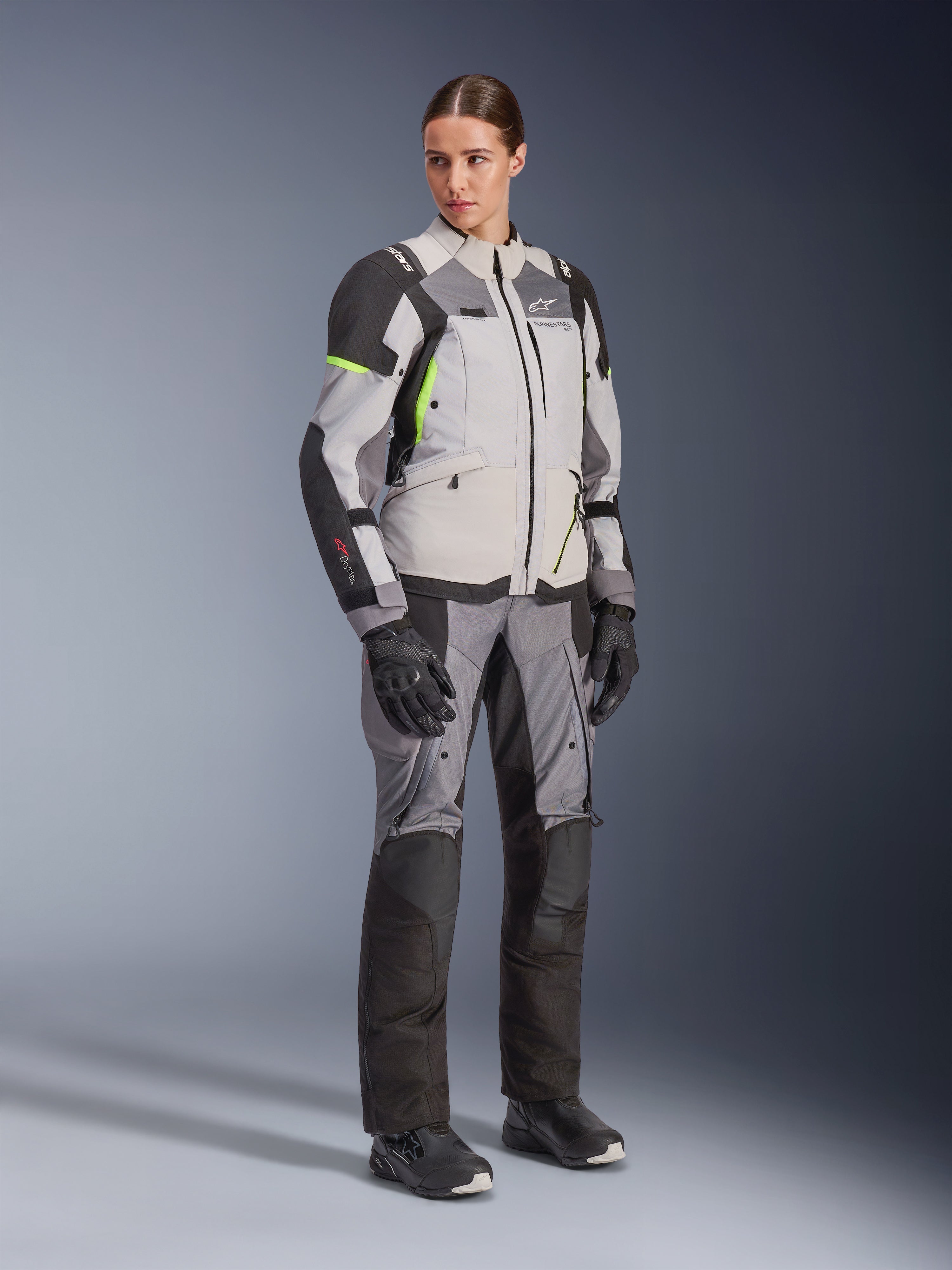 Mulher com casaco Alpinestars Stella Andes V4 Drystar® e calças a condizer, em Ice Gray e Dark Gray com detalhes em amarelo néon, conjunto completo de equipamento de aventura touring com luvas e botas pretas, fotografia de estúdio de corpo inteiro.