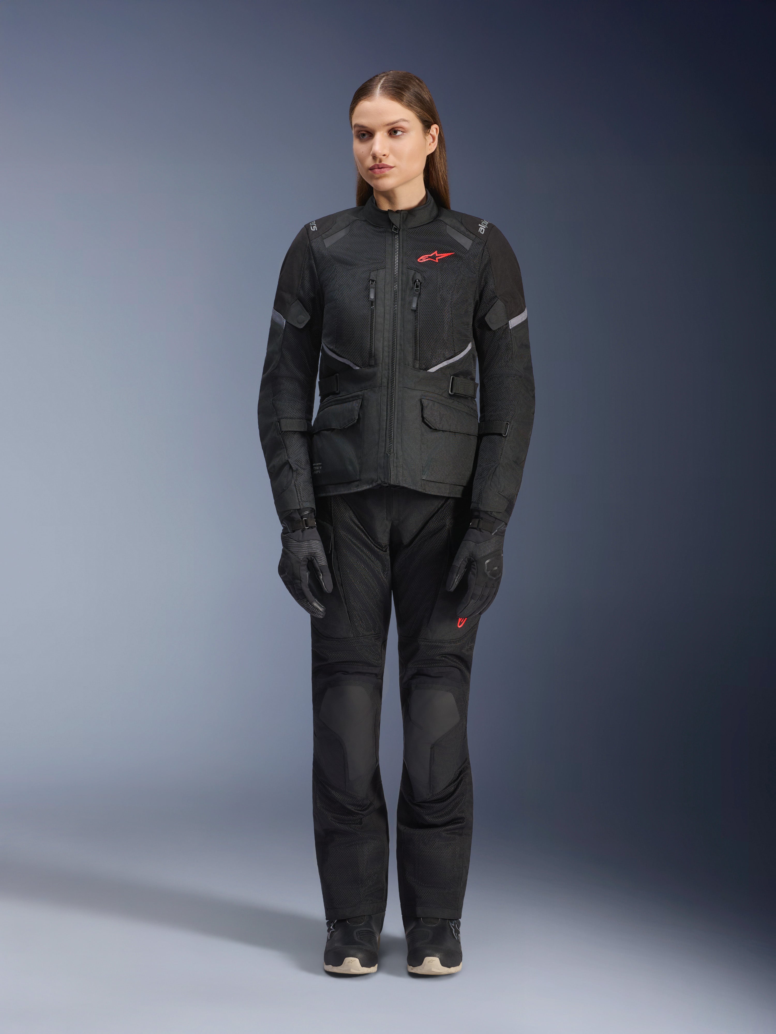 Modelo feminina a usar o casaco Alpinestars Stella Andes Air Drystar® em preto, combinado com calças de turismo e luvas pretas a condizer, com grandes painéis de ventilação em malha, detalhes do logótipo em vermelho e elementos refletores, fotografia profissional de estúdio de equipamento de motociclismo para mulher.