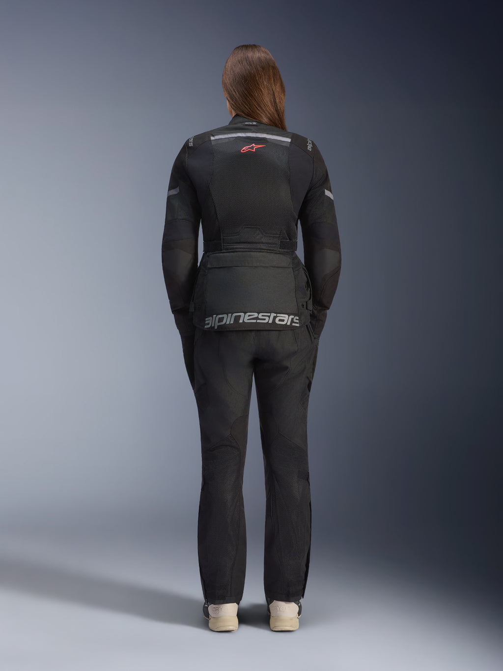 Mulher a usar o Casaco Alpinestars Stella Andes Air Drystar® e calças a condizer, preto, vista traseira exibindo a grande marca Alpinestars a cinzento na bolsa lombar, logótipo pequeno de estrela vermelha na parte superior das costas, painéis de ventilação em malha e detalhes refletores para adventure touring