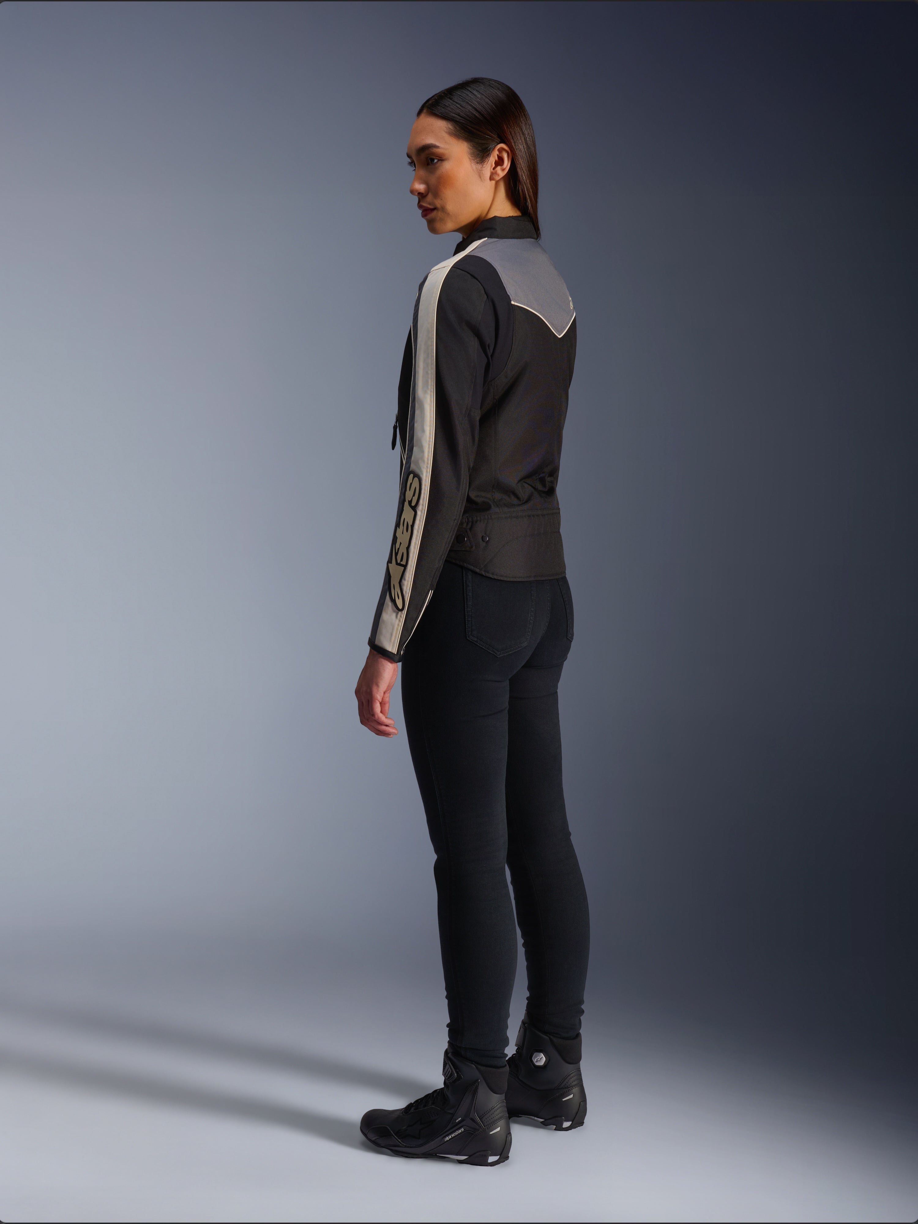 Modelo feminina vista de lado e de costas a usar o Casaco Alpinestars Stella T-Dyno WR em Preto Cinzento Escuro Ecru, com um design de inspiração vintage, riscas ecru nas mangas e um painel traseiro cinzento, combinado com calças pretas e calçado de motociclismo preto.