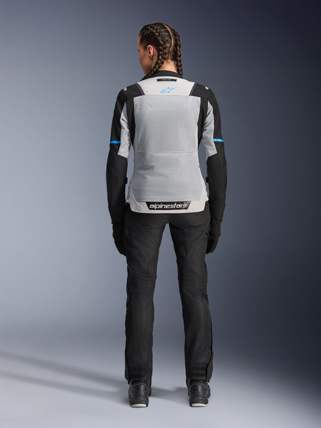 Alpinestars Stella ST-2 Air Jacket, Ice Gray Black, rear view on female model, featuring a large light gray mesh back panel, black sleeves with blue stripe accents, Alpinestars branding on the lower back, paired with black technical riding pants and boots, summer motorcycle touring gear