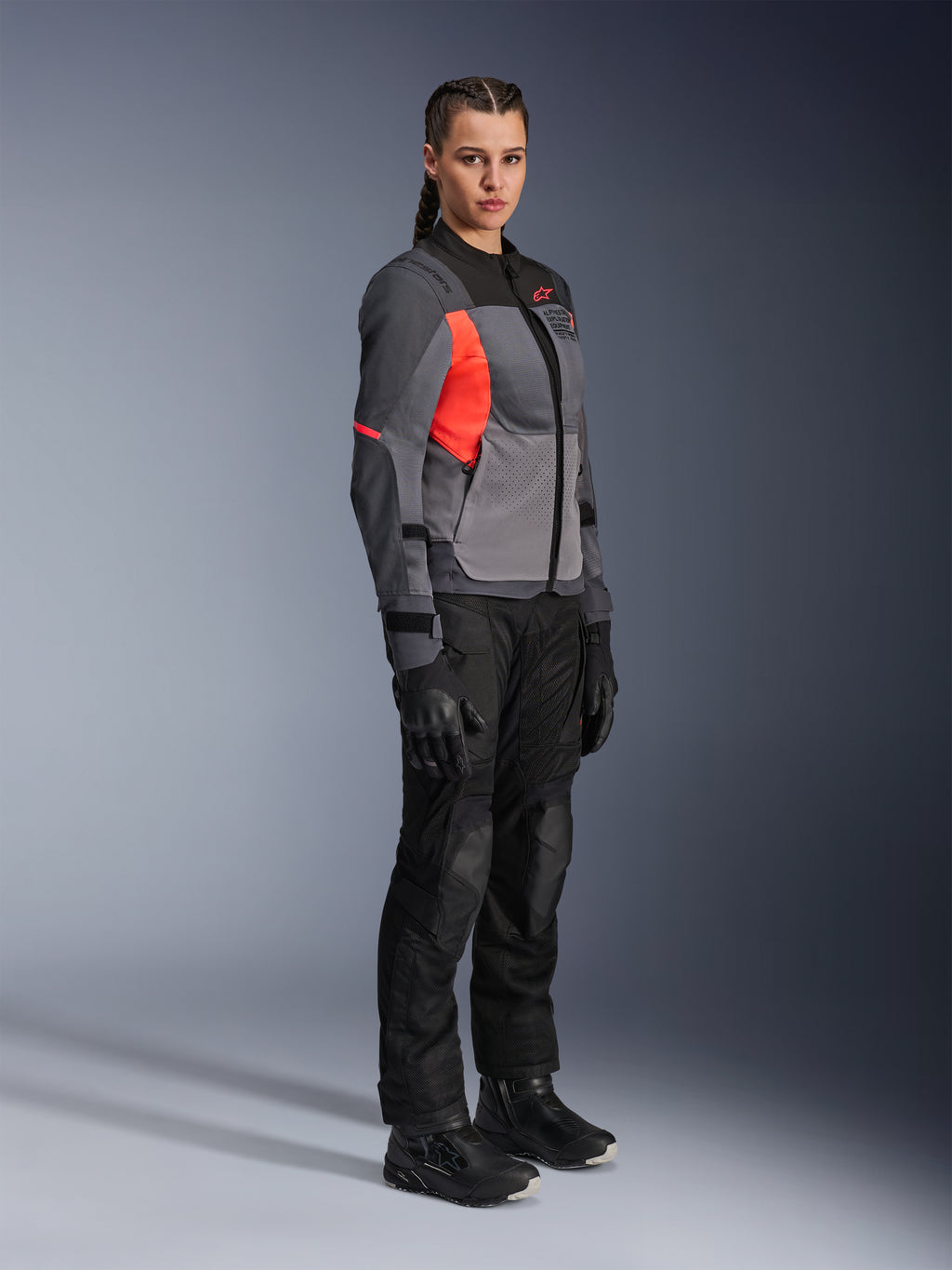Motociclista a usar o Casaco Alpinestars Stella ST-2 Air em cinzento escuro, cinzento e coral fluorescente, combinado com calças, luvas e botas de motociclismo pretas, apresentando um design touring moderno com painéis de malha e detalhes refletores.