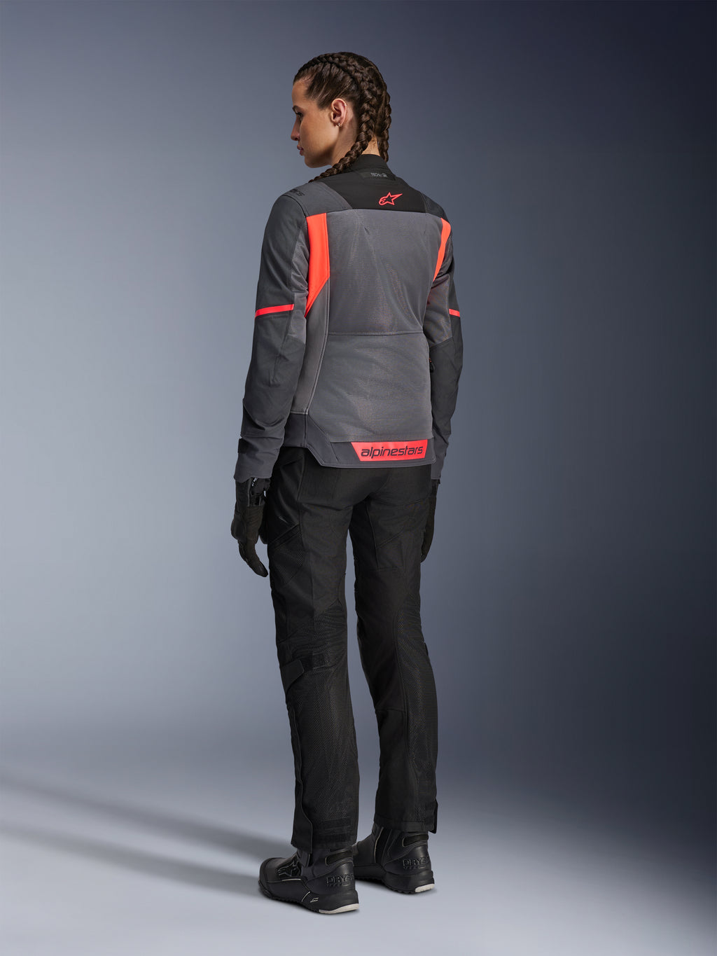 Mulher a usar o Casaco Alpinestars Stella St-2 Air, casaco de touring para motociclismo, cinzento profundo e cinzento escuro com detalhes em coral fluo, combinado com calças e luvas de motociclismo pretas, vista traseira mostrando amplos painéis de malha e marca refletora em coral, concebido para aventuras de condução no verão
