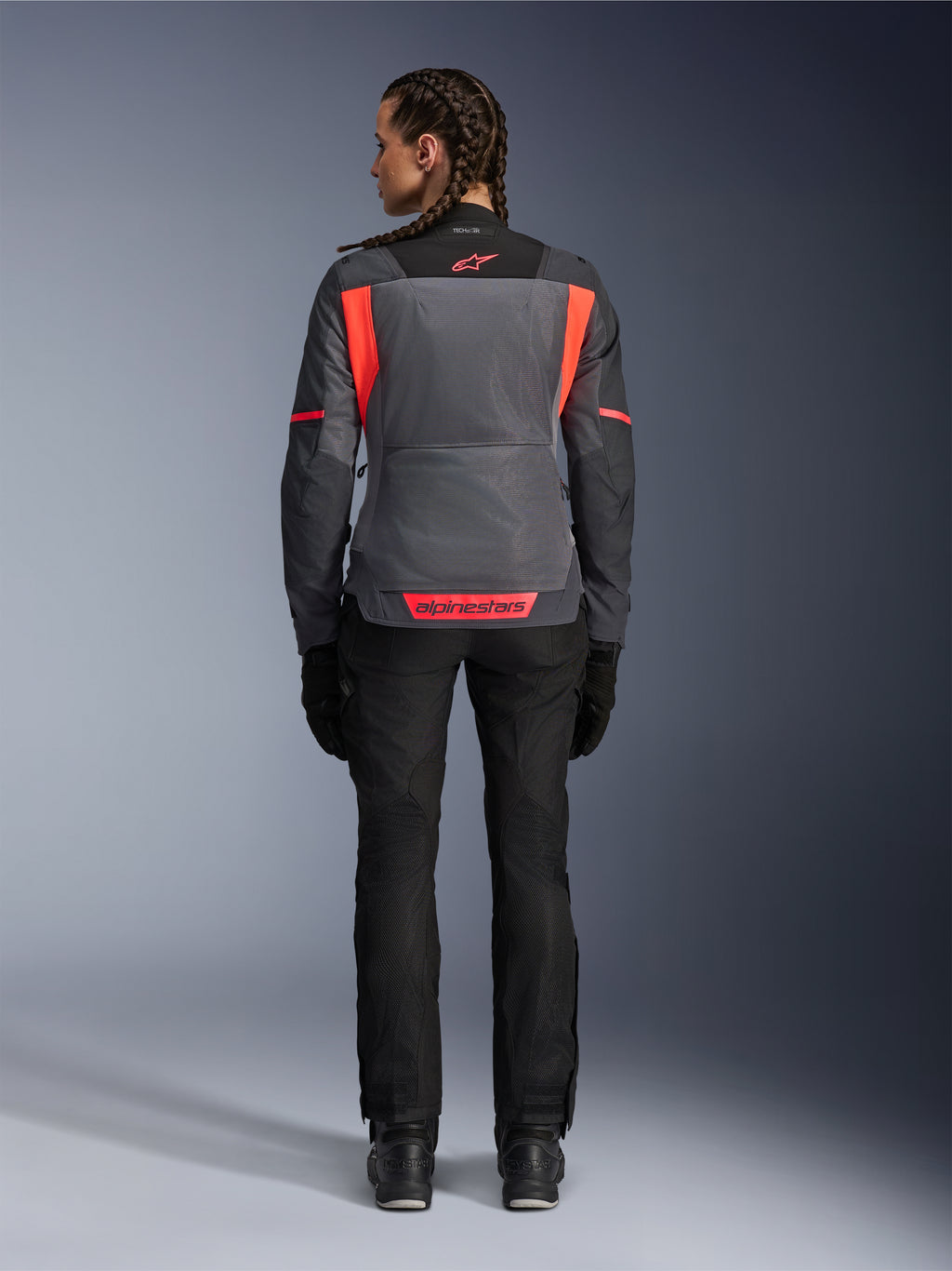 Vista traseira de uma modelo a usar o Casaco Alpinestars Stella St-2 Air em Cinzento Profundo e Cinzento Escuro com detalhes Coral Fluo, apresentando grandes painéis de ventilação em malha nas costas e pormenores de logótipo refletores, combinada com calças e luvas de motociclismo pretas.