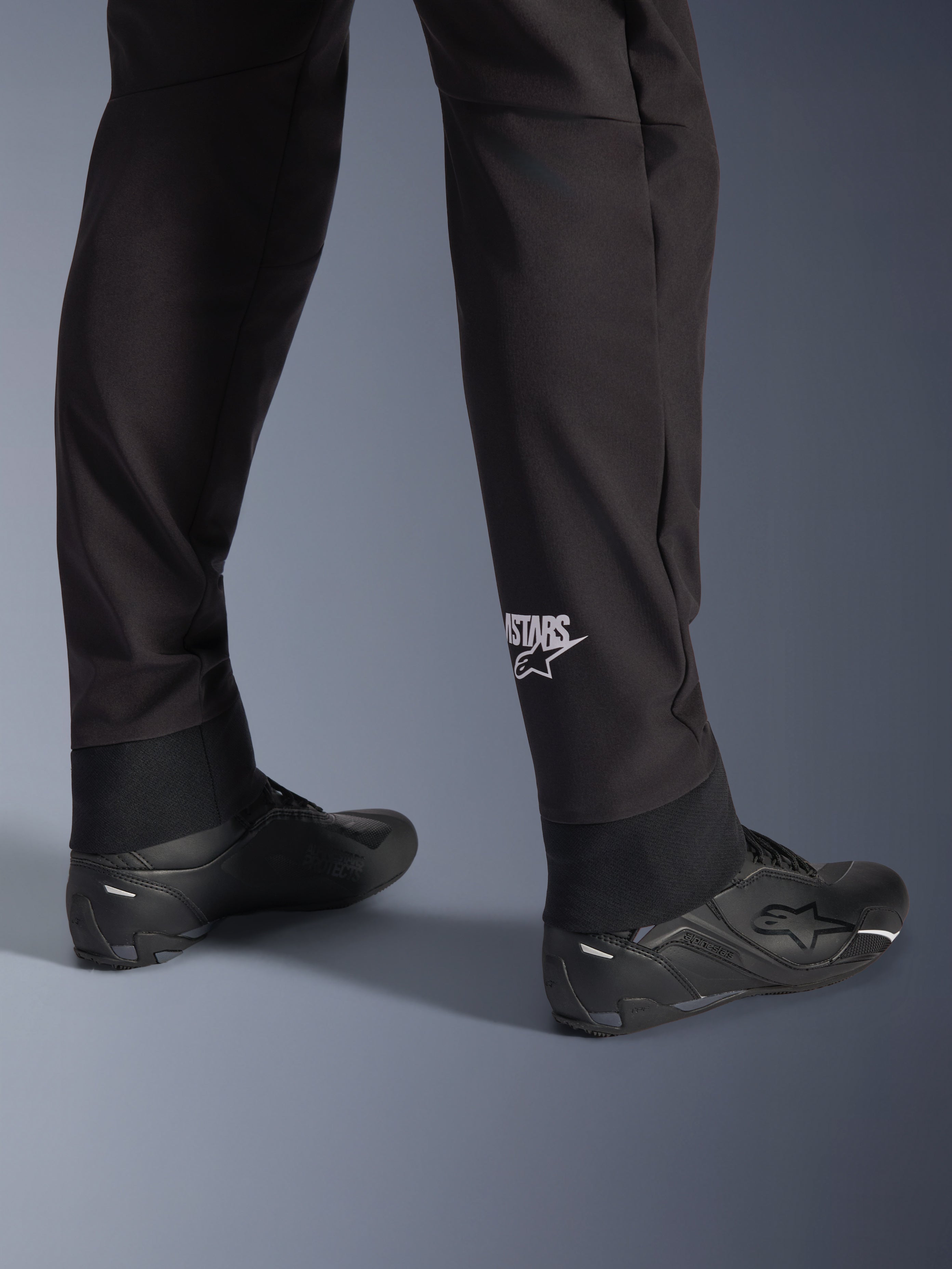 Alpinestars Aeroshell Pants, Black softshell motorcycle pants, close-up of lower leg and elasticated hem, white Alpinestars logo on calf, styled with black riding shoes, modern crossover sport design