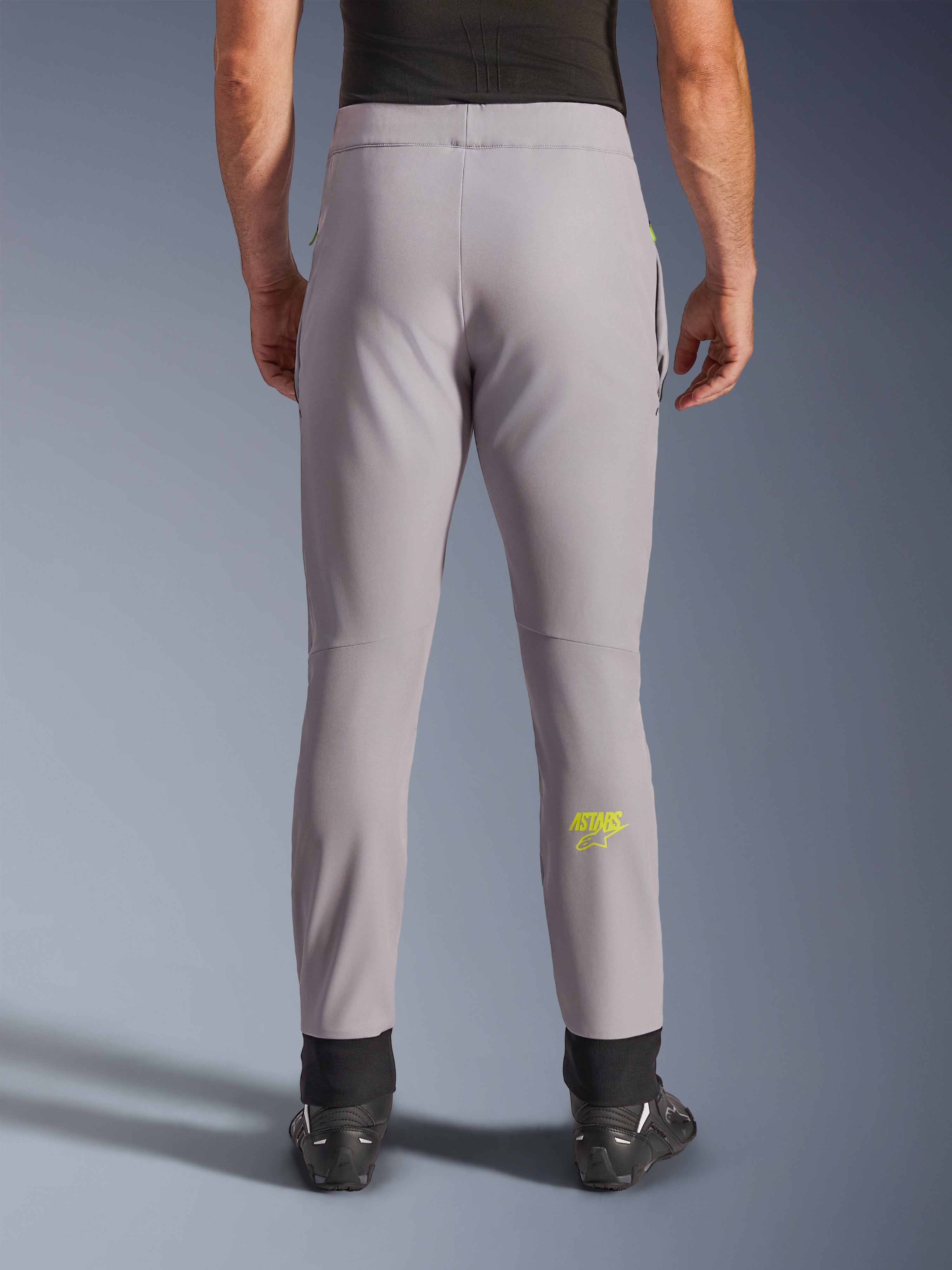 Alpinestars Aeroshell Pants, calças de motociclismo, cinzento médio e amarelo néon, vista traseira mostrando o logótipo Astars amarelo na barriga da perna, punhos elásticos pretos nos tornozelos, design têxtil softshell moderno para condução em estrada e casual