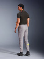 Calças Alpinestars Aeroshell, Cinzento Médio Amarelo Fluo, vista lateral traseira de modelo masculino a usar calças de motociclismo em softshell elástico, logótipo Astars amarelo na barriga da perna esquerda, combinadas com uma camada base preta e sapatos de condução para uma silhueta desportiva moderna.