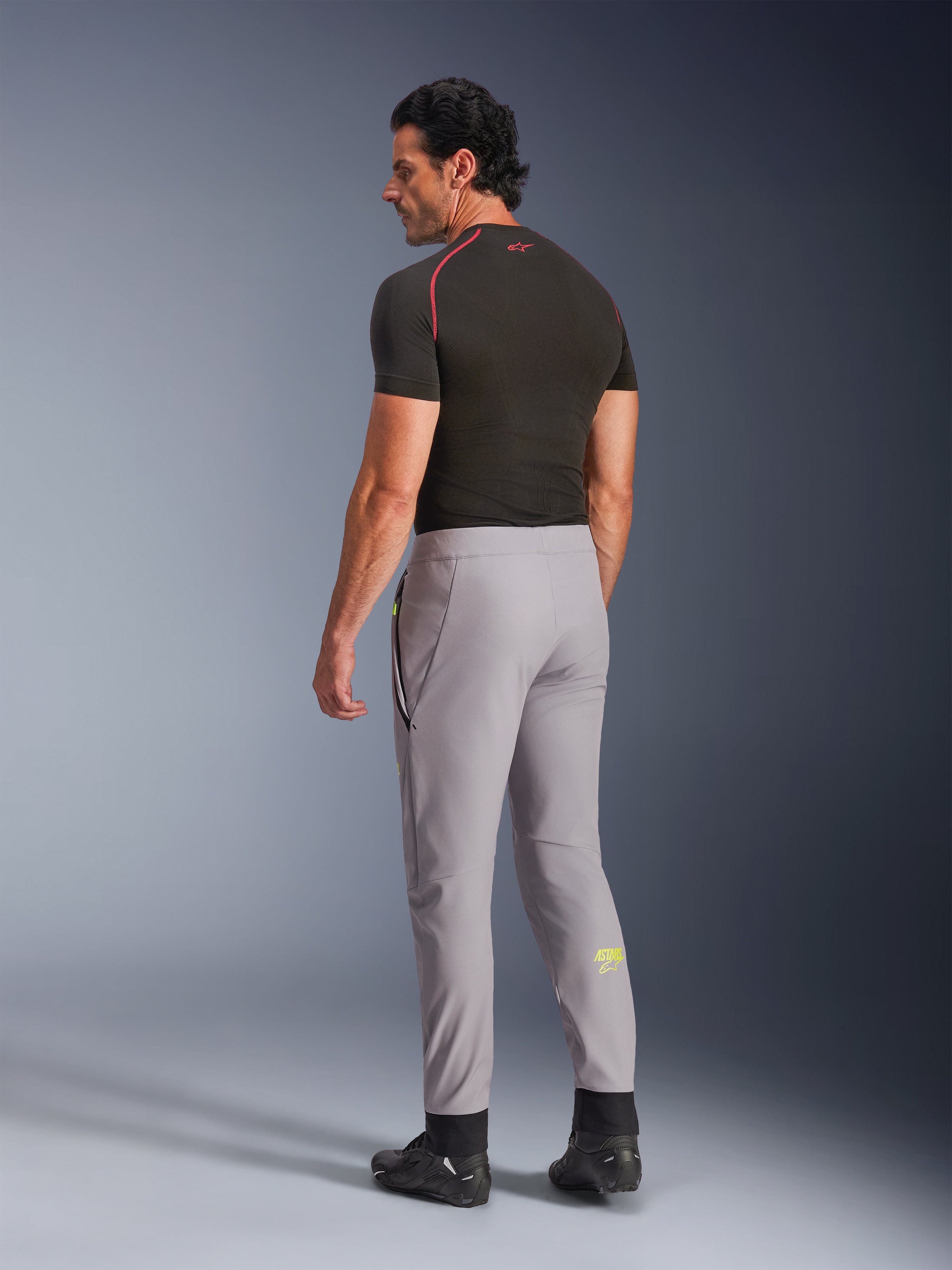 Calças Alpinestars Aeroshell, Cinzento Médio Amarelo Fluo, vista lateral traseira de modelo masculino a usar calças de motociclismo em softshell elástico, logótipo Astars amarelo na barriga da perna esquerda, combinadas com uma camada base preta e sapatos de condução para uma silhueta desportiva moderna.