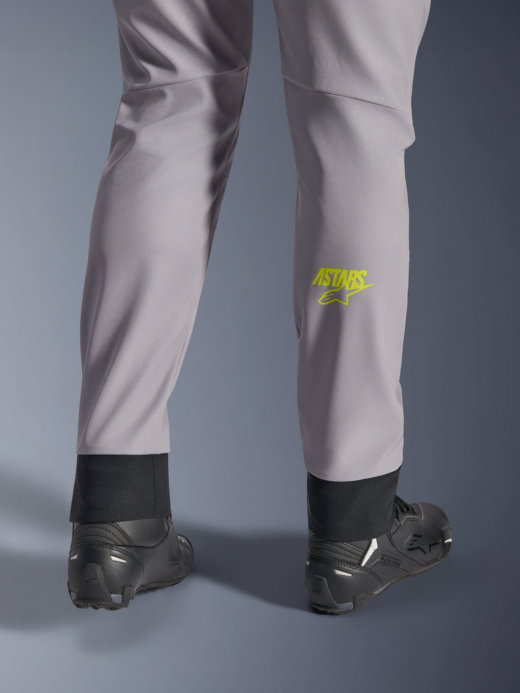 Calças Alpinestars Aeroshell, cinzento médio com logótipo Astars em amarelo fluo na barriga da perna, vistas de trás com calçado de motociclismo preto, apresentando uma bainha inferior elástica e uma silhueta afunilada moderna