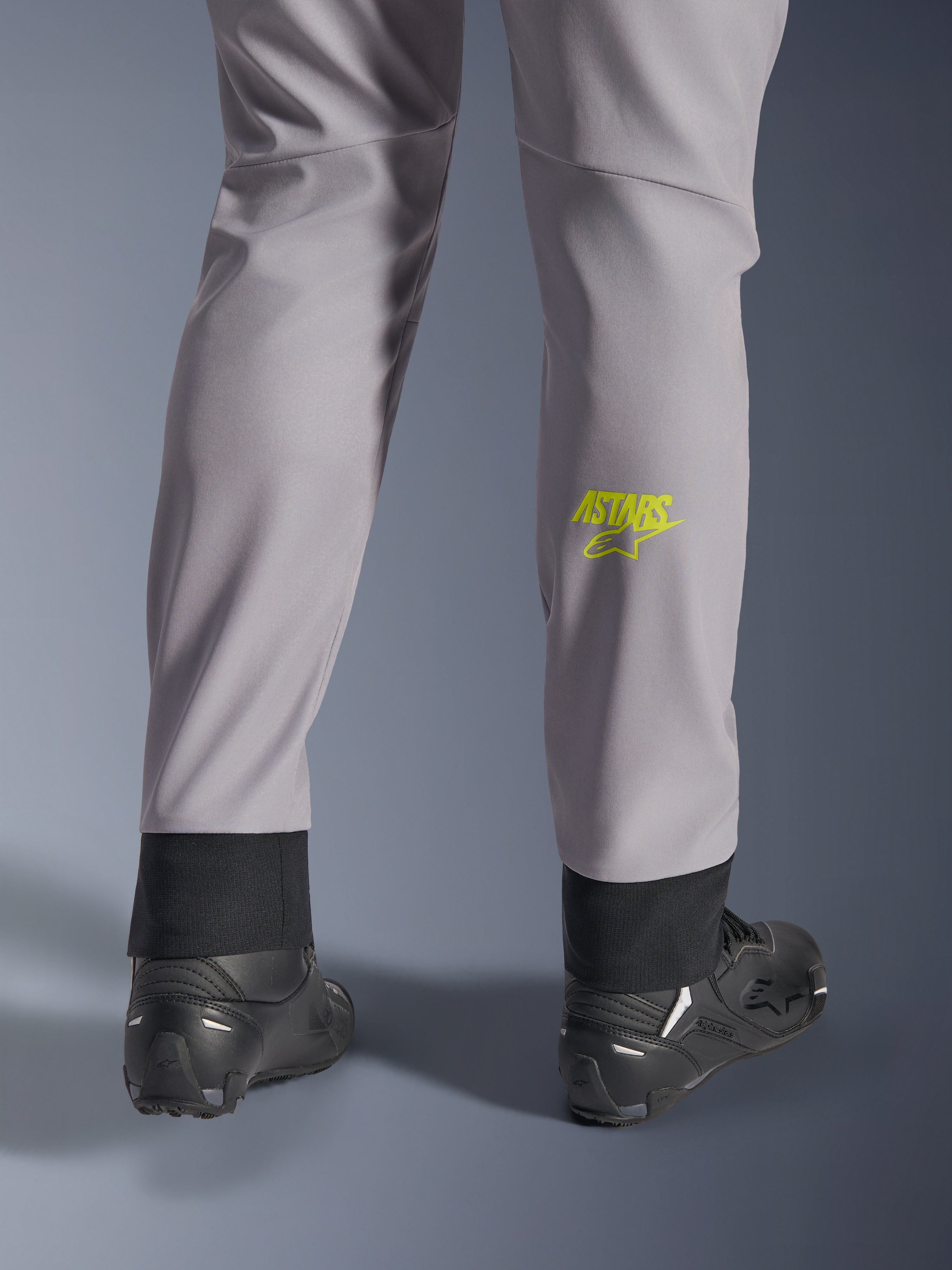 Calças Alpinestars Aeroshell, cinzento médio com logótipo Astars em amarelo fluo na barriga da perna, vistas de trás com calçado de motociclismo preto, apresentando uma bainha inferior elástica e uma silhueta afunilada moderna
