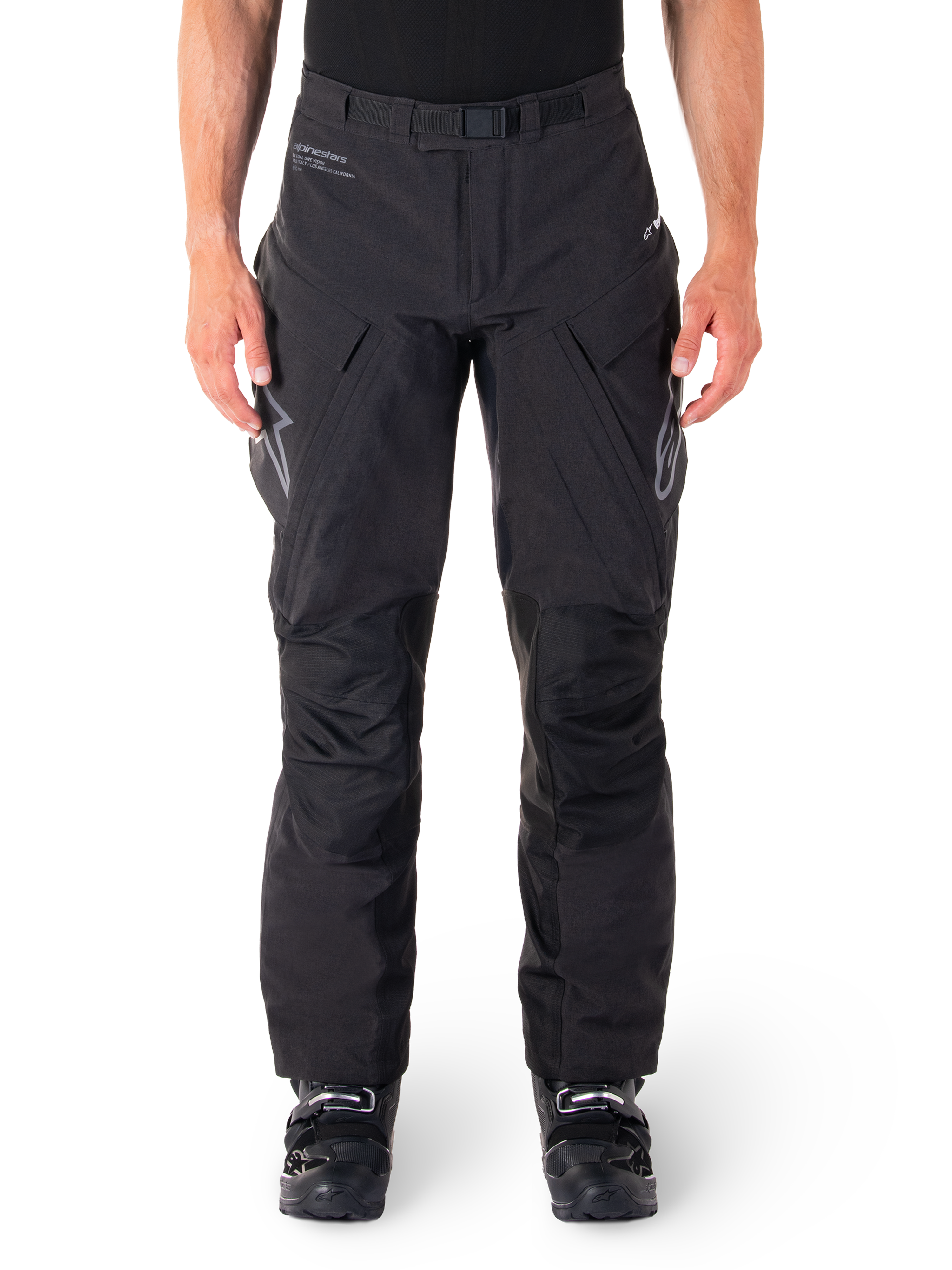 Alpinestars Hyde XT Drystar Pants, calças de aventura e turismo pretas, vestidas por um piloto com botas pretas, com logótipos cinzentos nas coxas, cinto integrado, painéis de joelho reforçados e construção têxtil impermeável